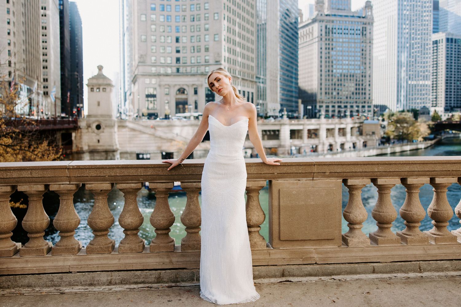Bride in elegant white dress posing on stone balcony with Chicago cityscape in background.