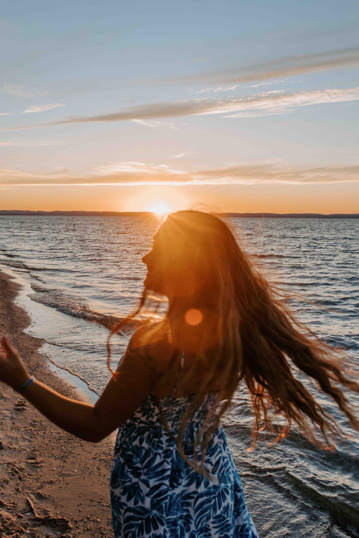 Figure with windswept hair twirls in dress against golden sunset on beach.