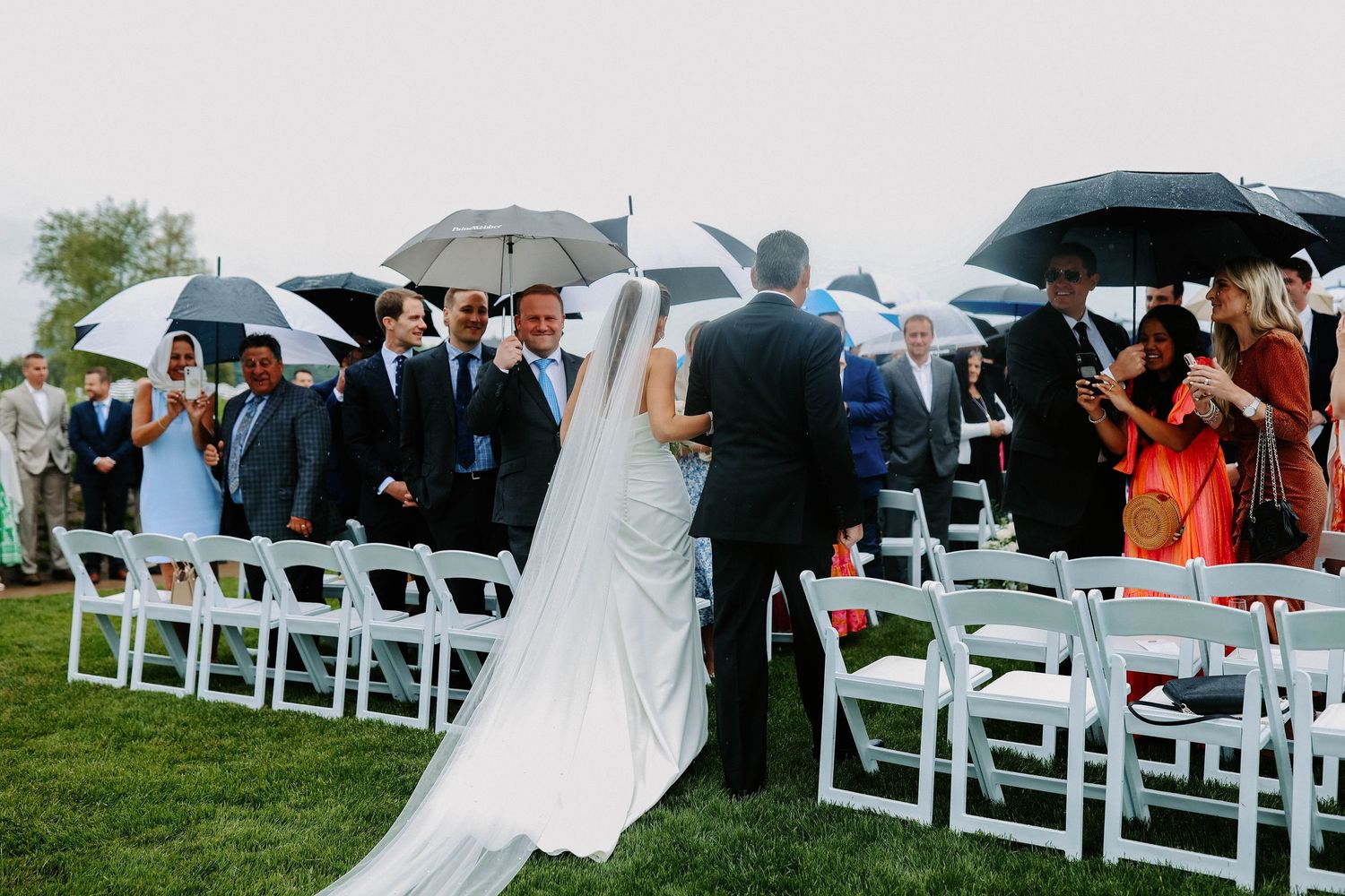 Guests with umbrellas seated in white chairs at an outdoor waterfront wedding ceremony.