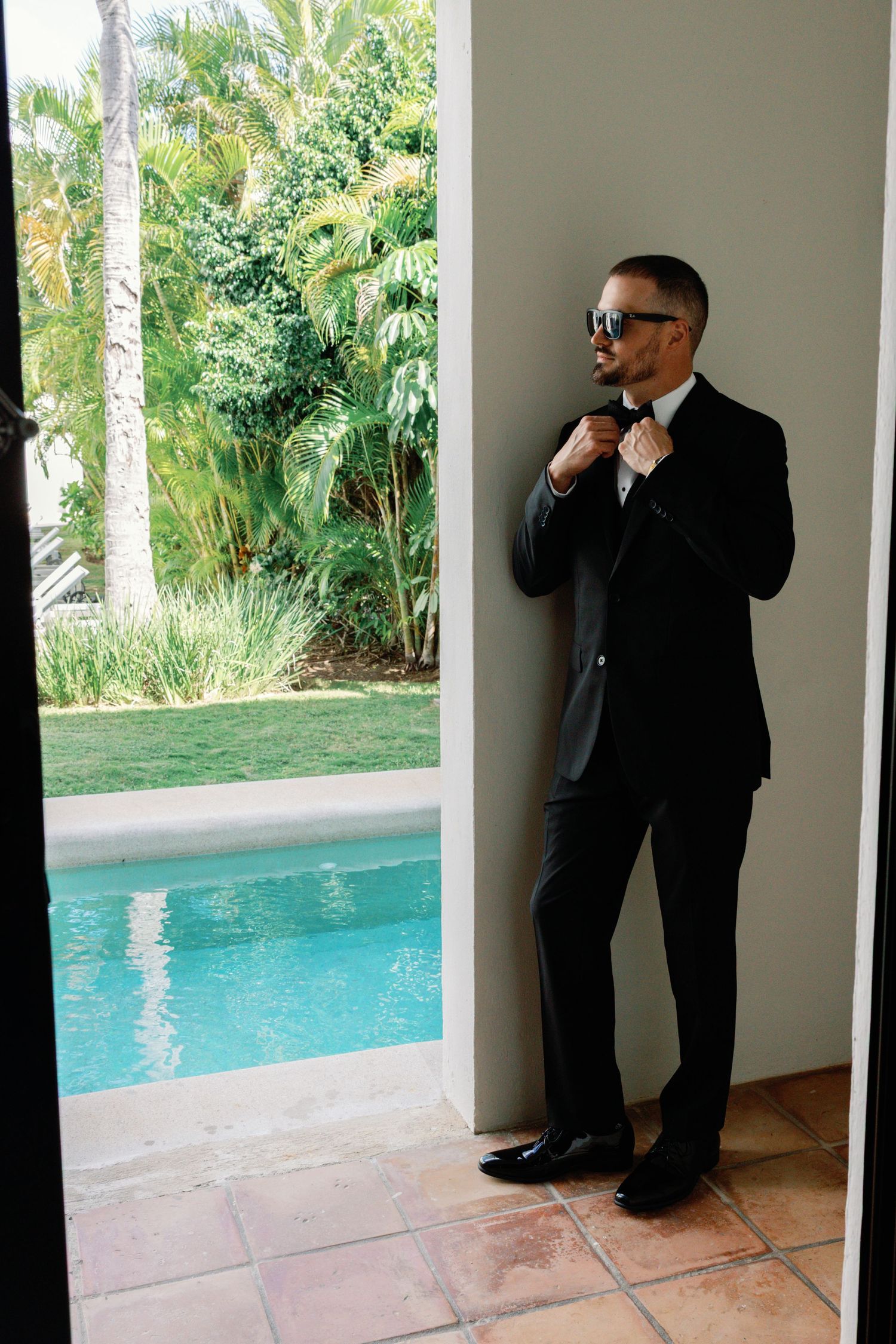A formal suited figure adjusts their tie while standing near a swimming pool surrounded by tropical palm trees.