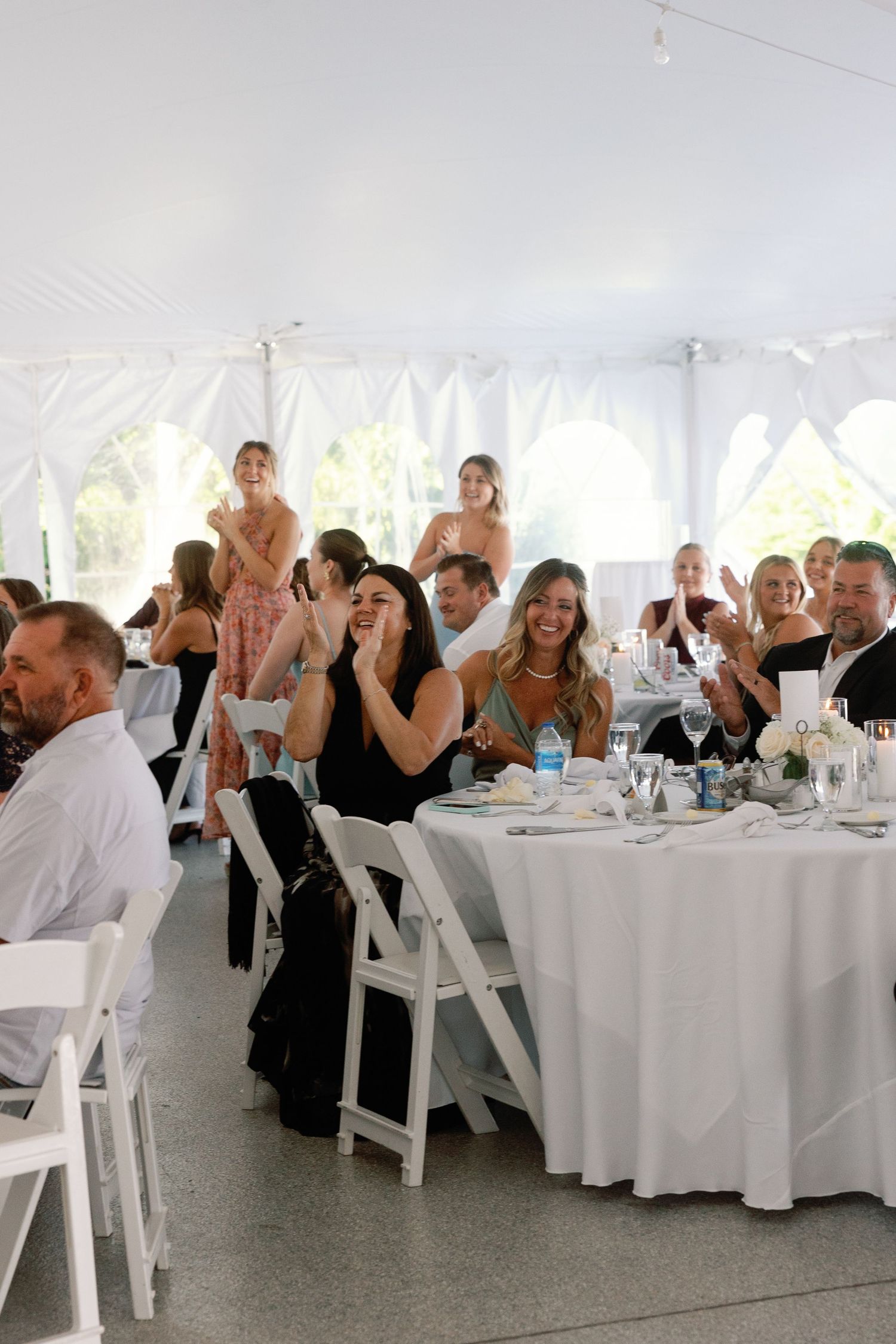 Wedding reception guests celebrate with raised hands at tables under a white tent.