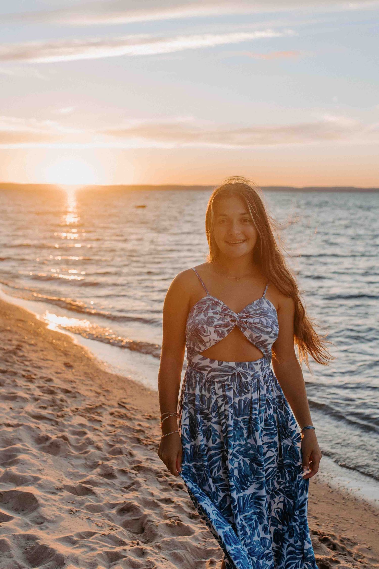 Woman wearing blue patterned dress walks along sandy shoreline during vibrant sunset.