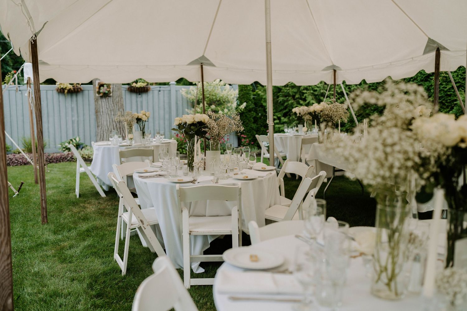 Elegant outdoor wedding reception setup with white tables and chairs under a tent surrounded by greenery.