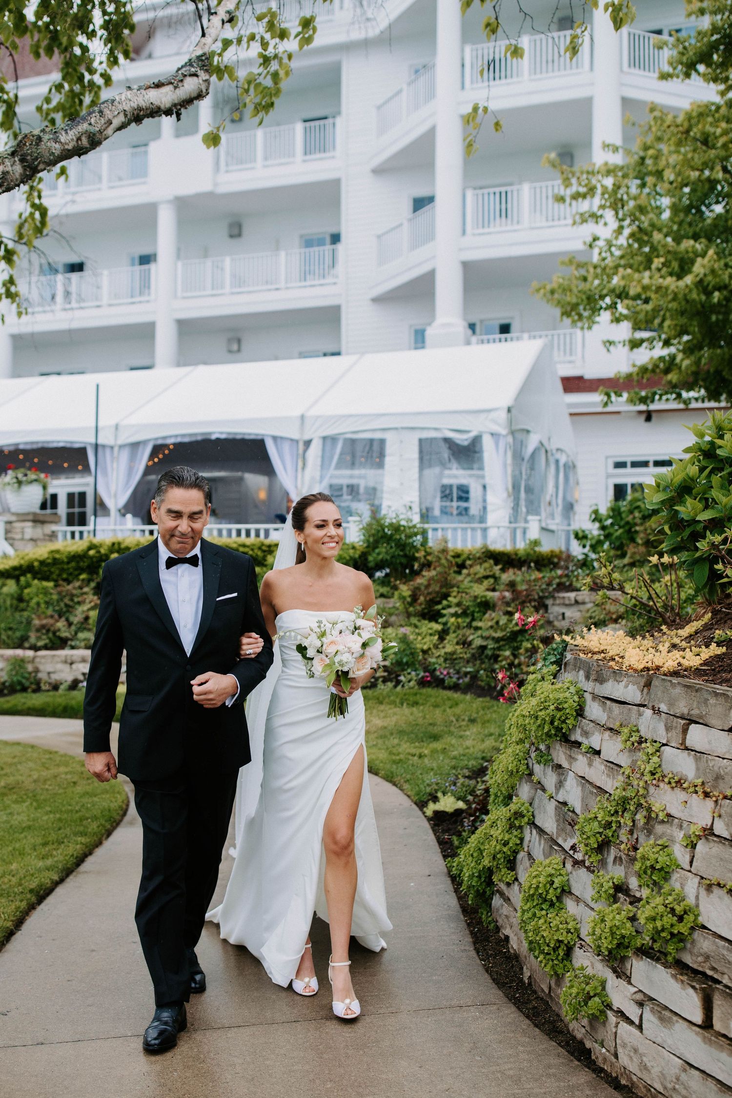 Wedding party members stroll along curved pathway beside stone wall and landscaped gardens.