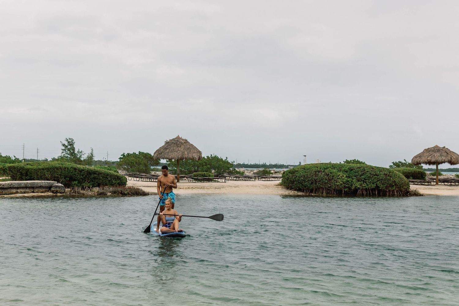Person paddleboarding near tropical shoreline with thatched huts in background.
