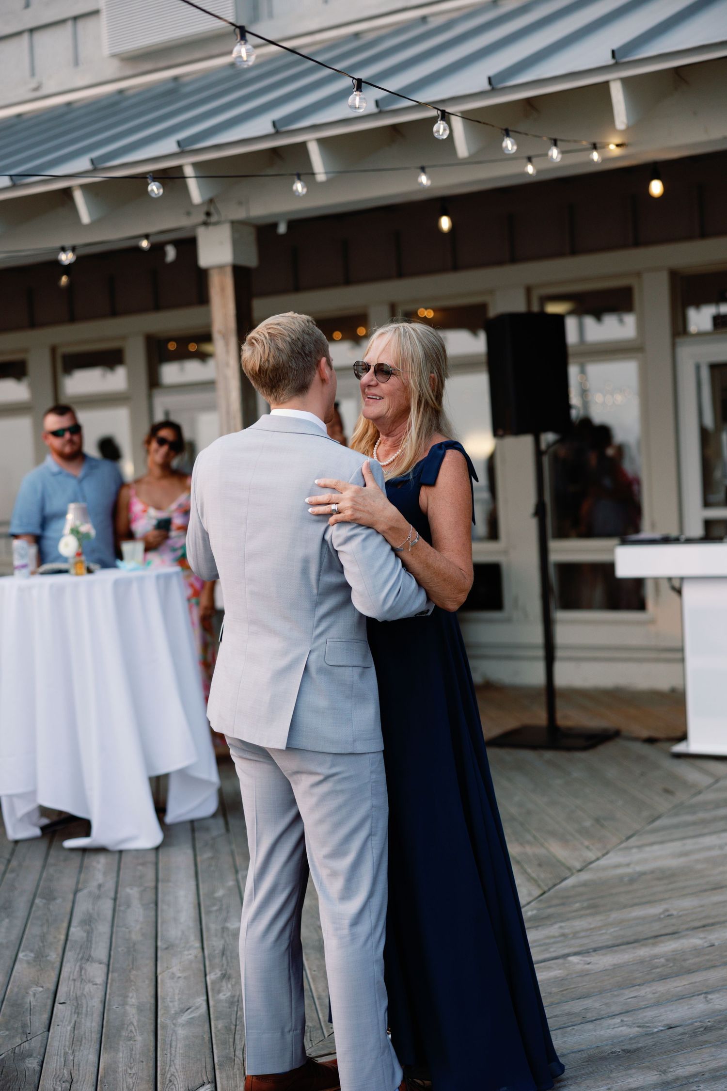 Wedding guests mingle on a covered outdoor deck with string lights.