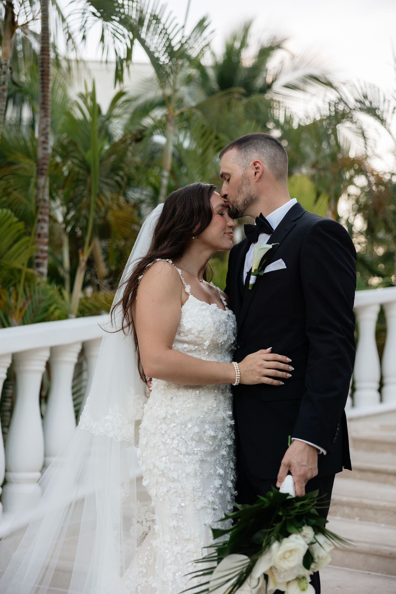 A romantic wedding couple shares intimate moments on a tropical terrace with white balustrades and palm trees.