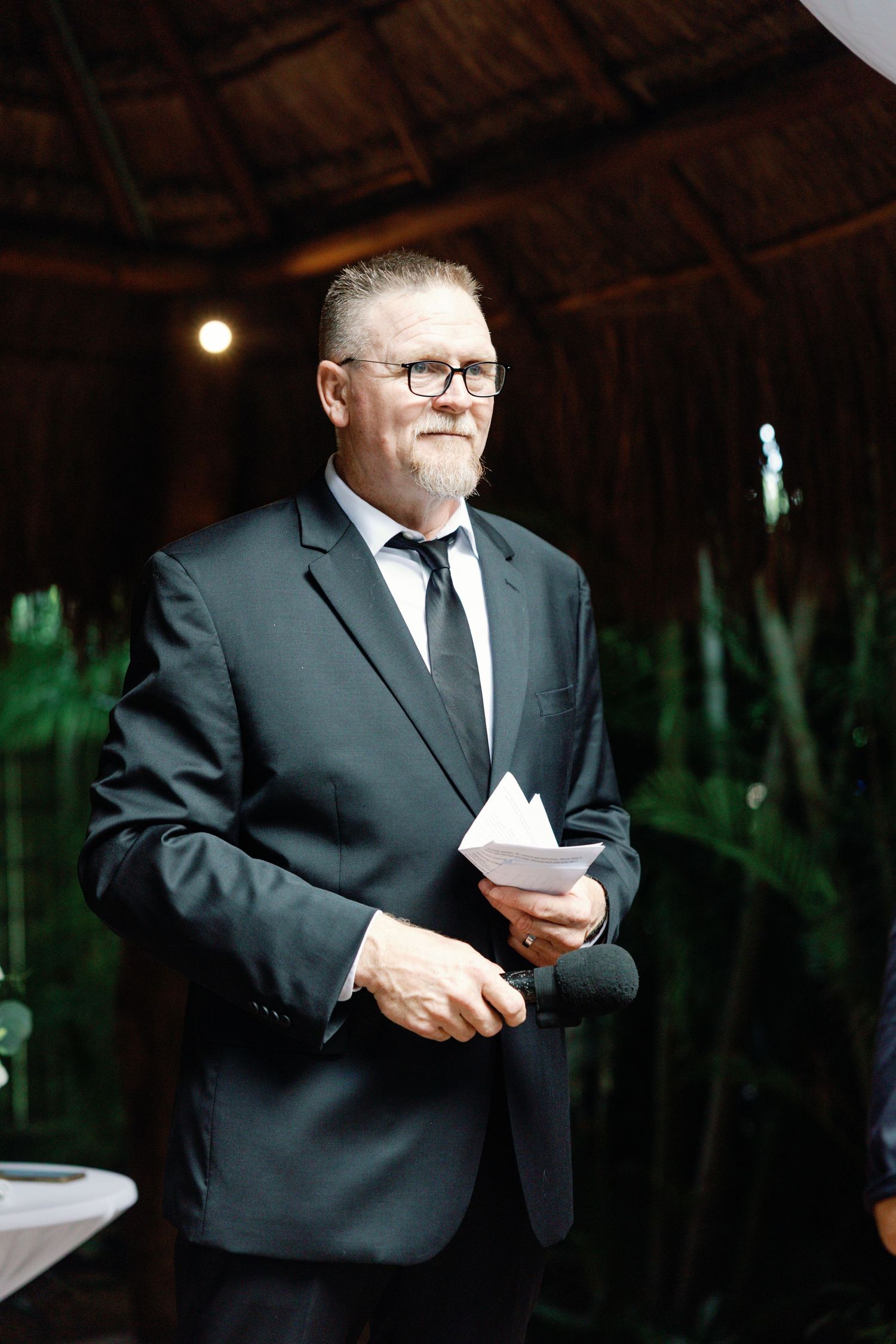 Groomsman in black suit reads from papers during wedding ceremony.