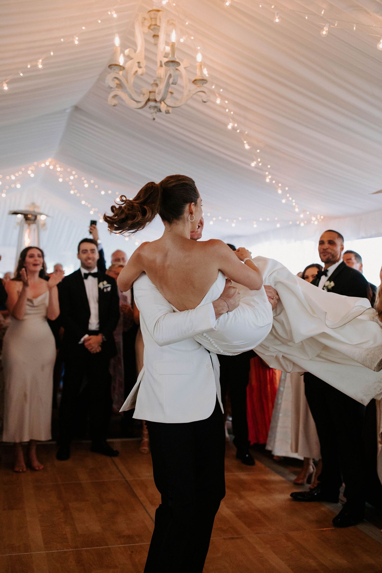 Wedding couples dancing energetically under white tent with string lights and chandeliers.