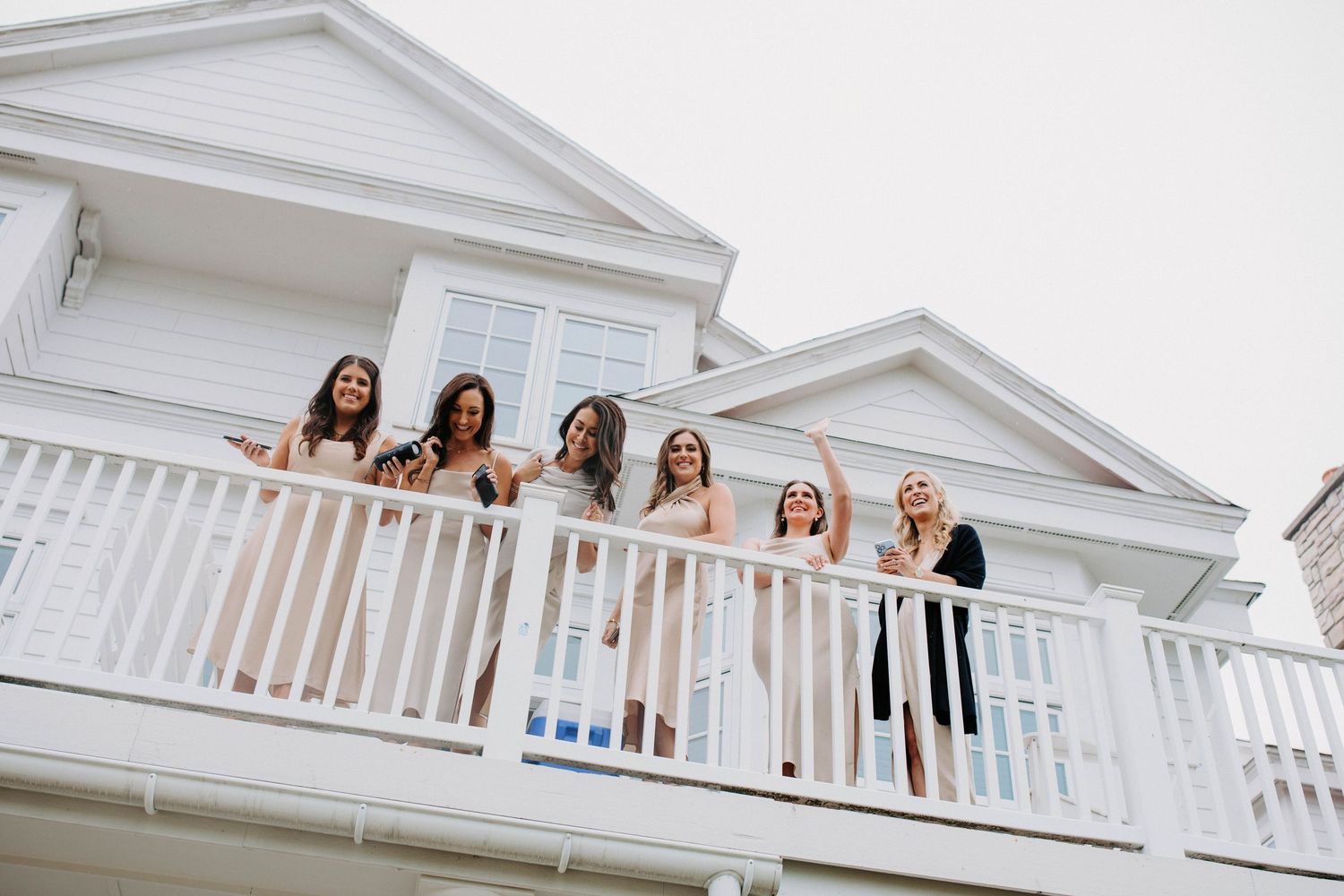 Bridesmaids in light-colored dresses smiling from a white balcony railing of an elegant coastal home.