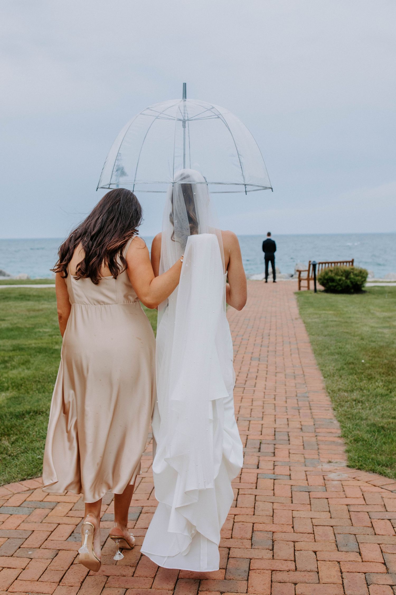 Two people walk down a brick pathway with clear umbrellas towards the ocean.