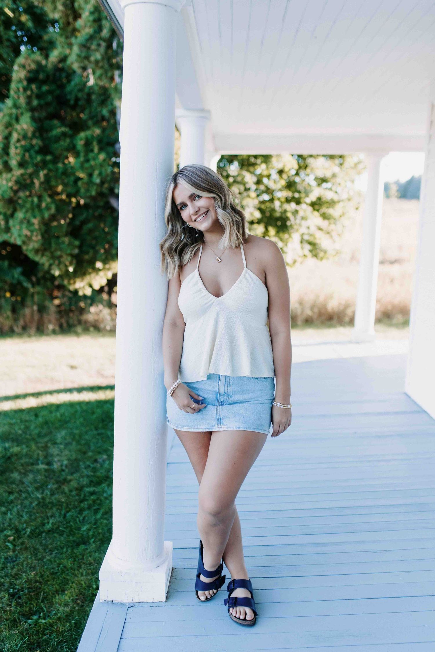Person in white top and denim shorts sits on white porch with greenery in background.