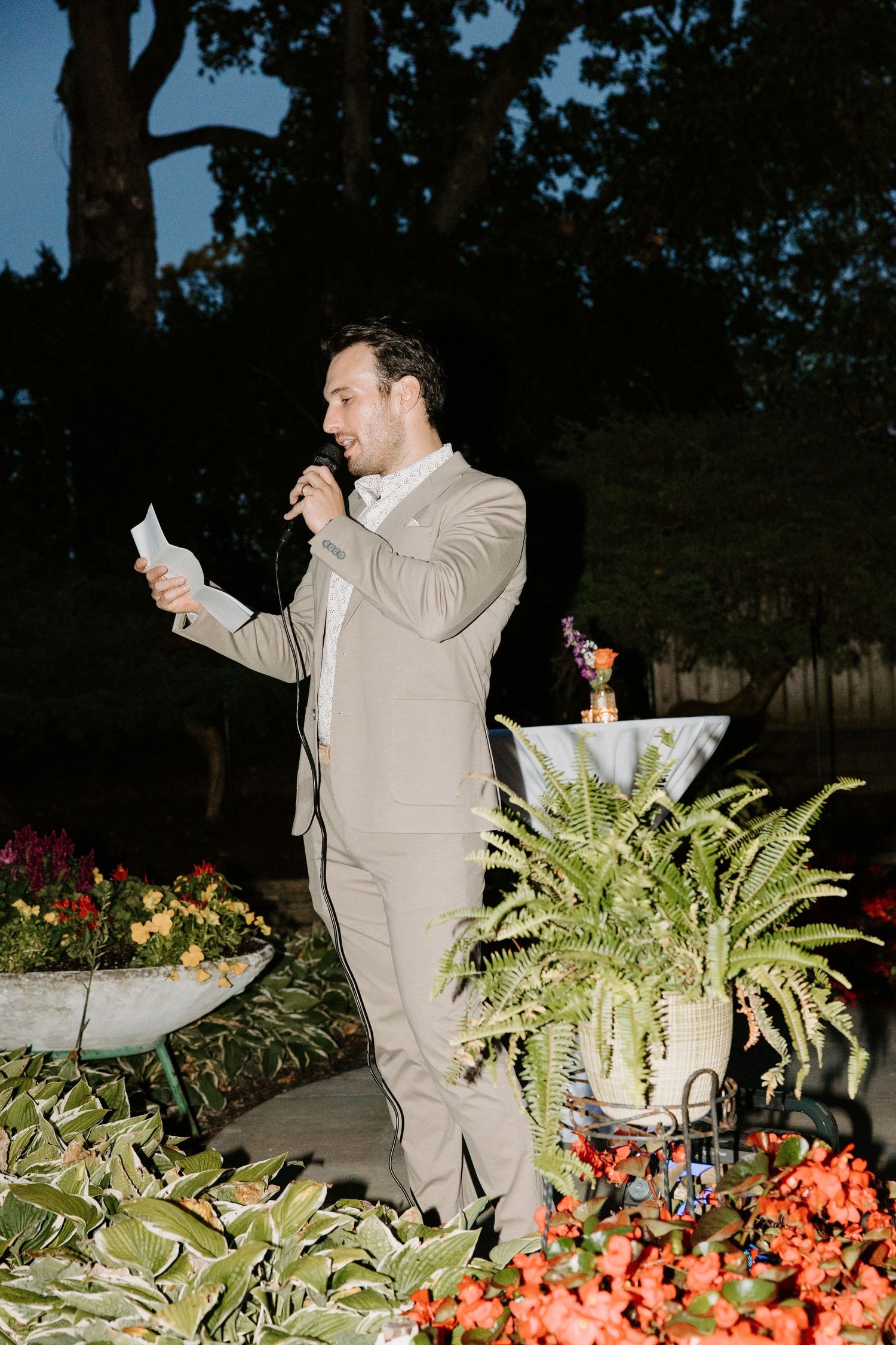 Person in a beige suit dances energetically among garden flowers at an outdoor evening event.