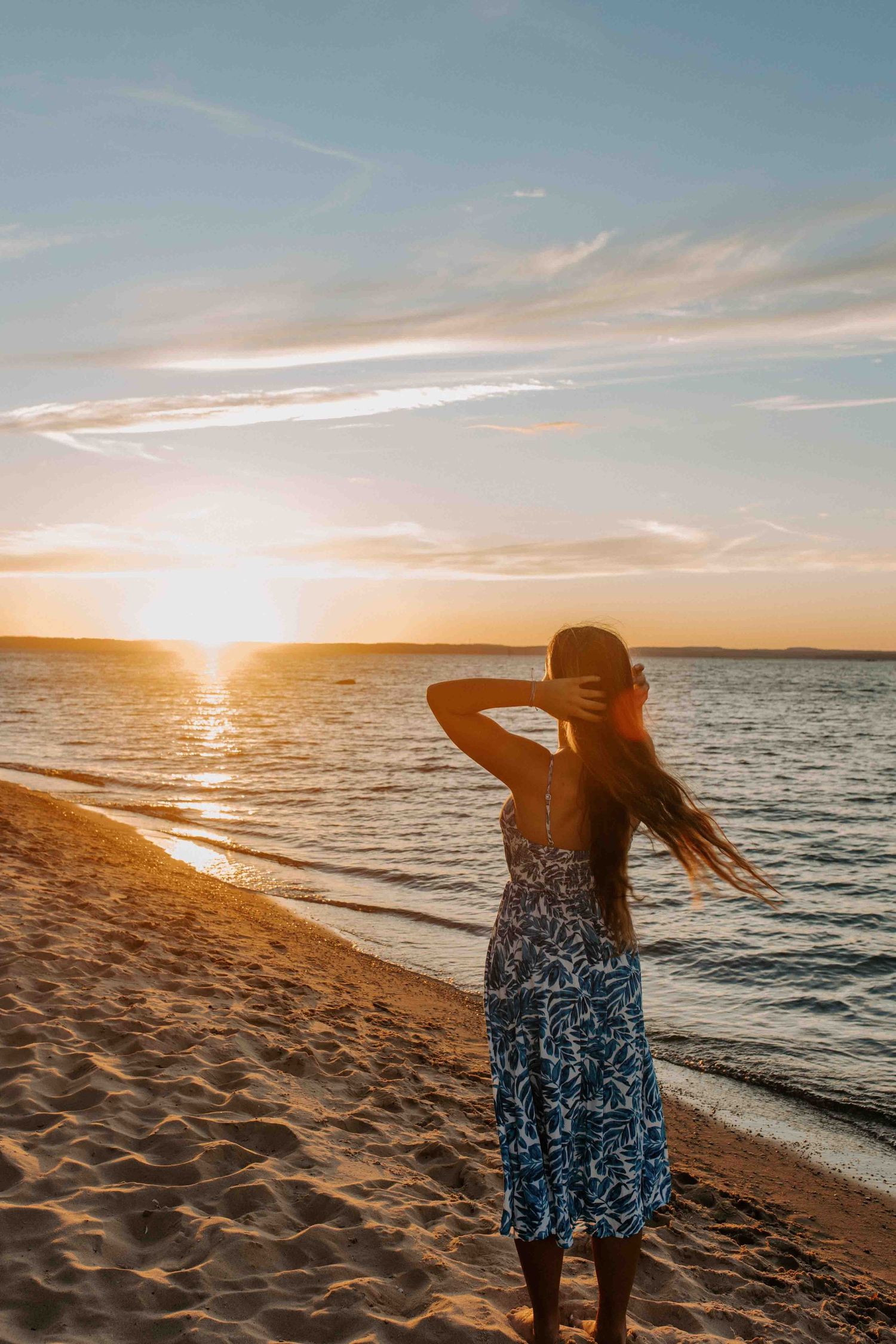 Person in a blue dress stands on beach watching sunset over calm waters.