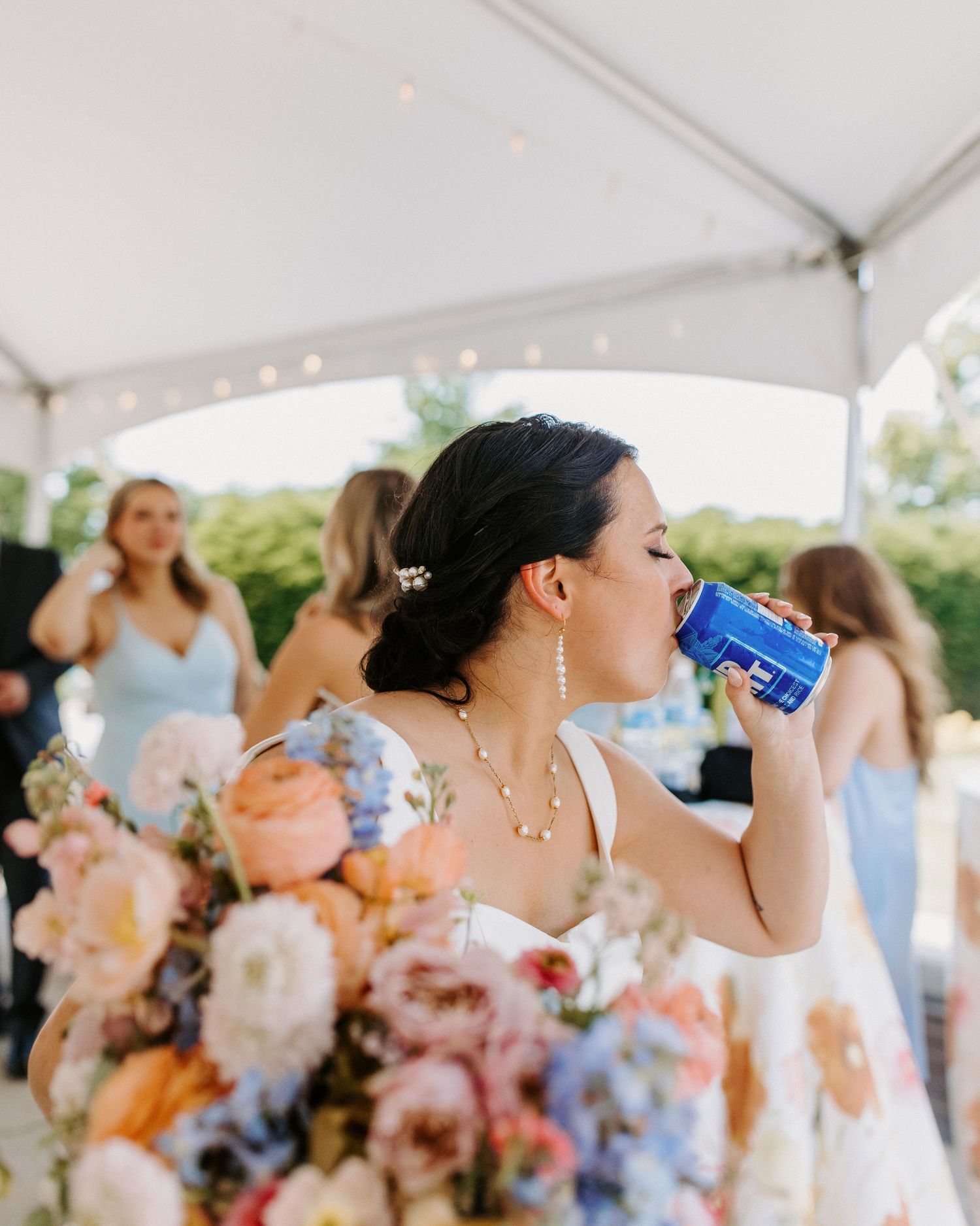 Wedding party guests enjoying drinks under a white tent at an outdoor celebration.