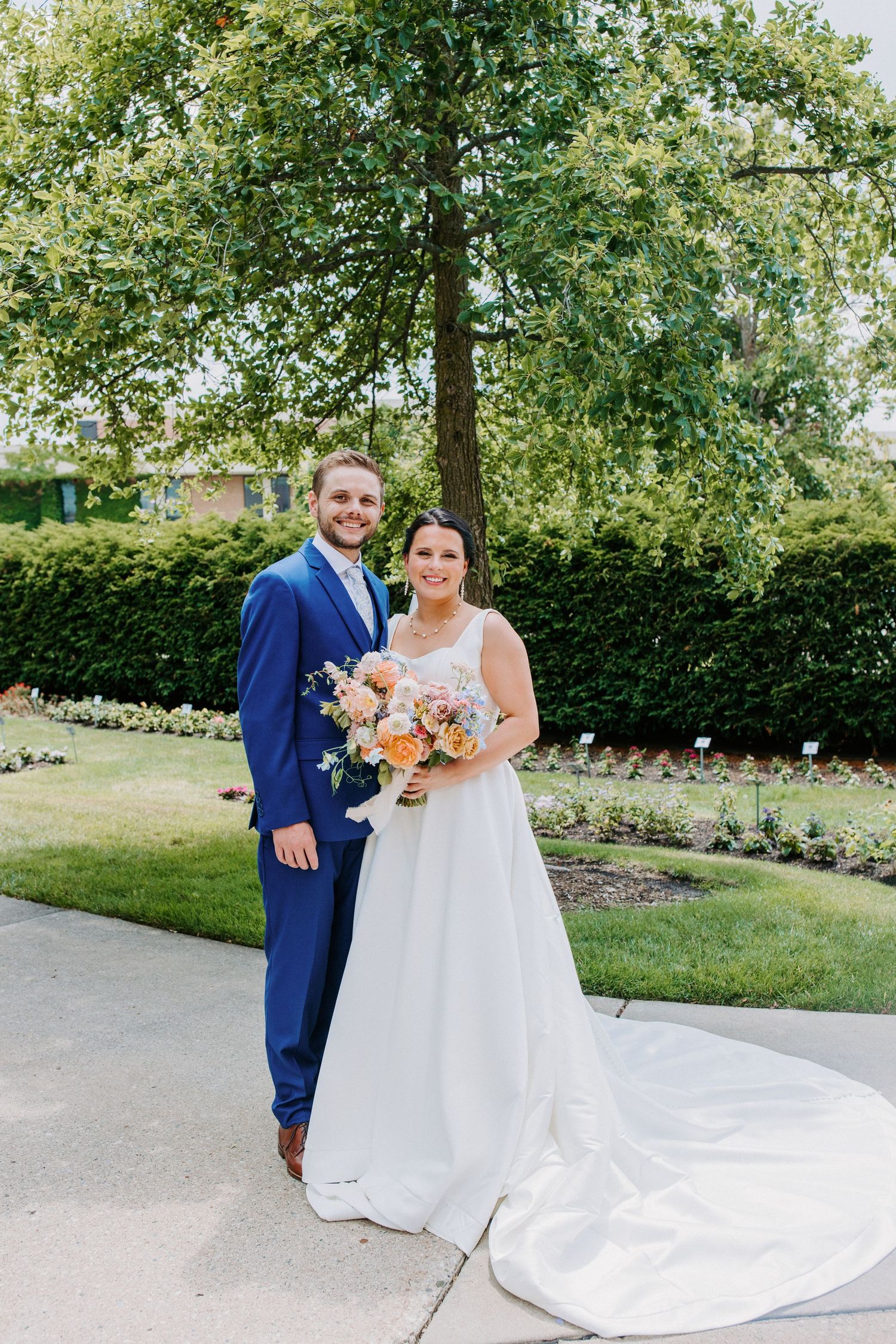 A wedding couple poses outdoors in front of lush greenery with the bride holding a colorful bouquet.