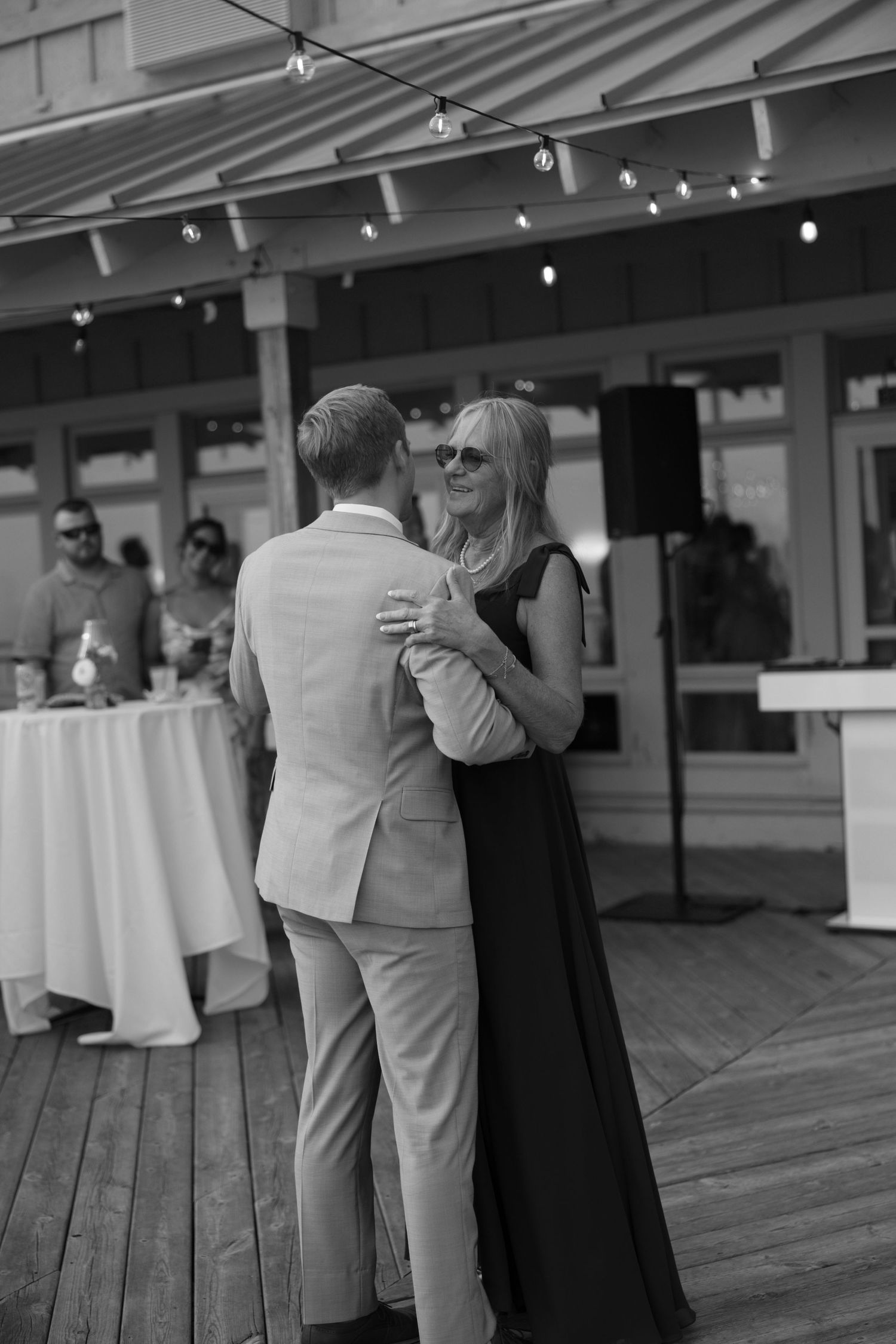 Wedding guests gather on a covered deck with string lights overhead.