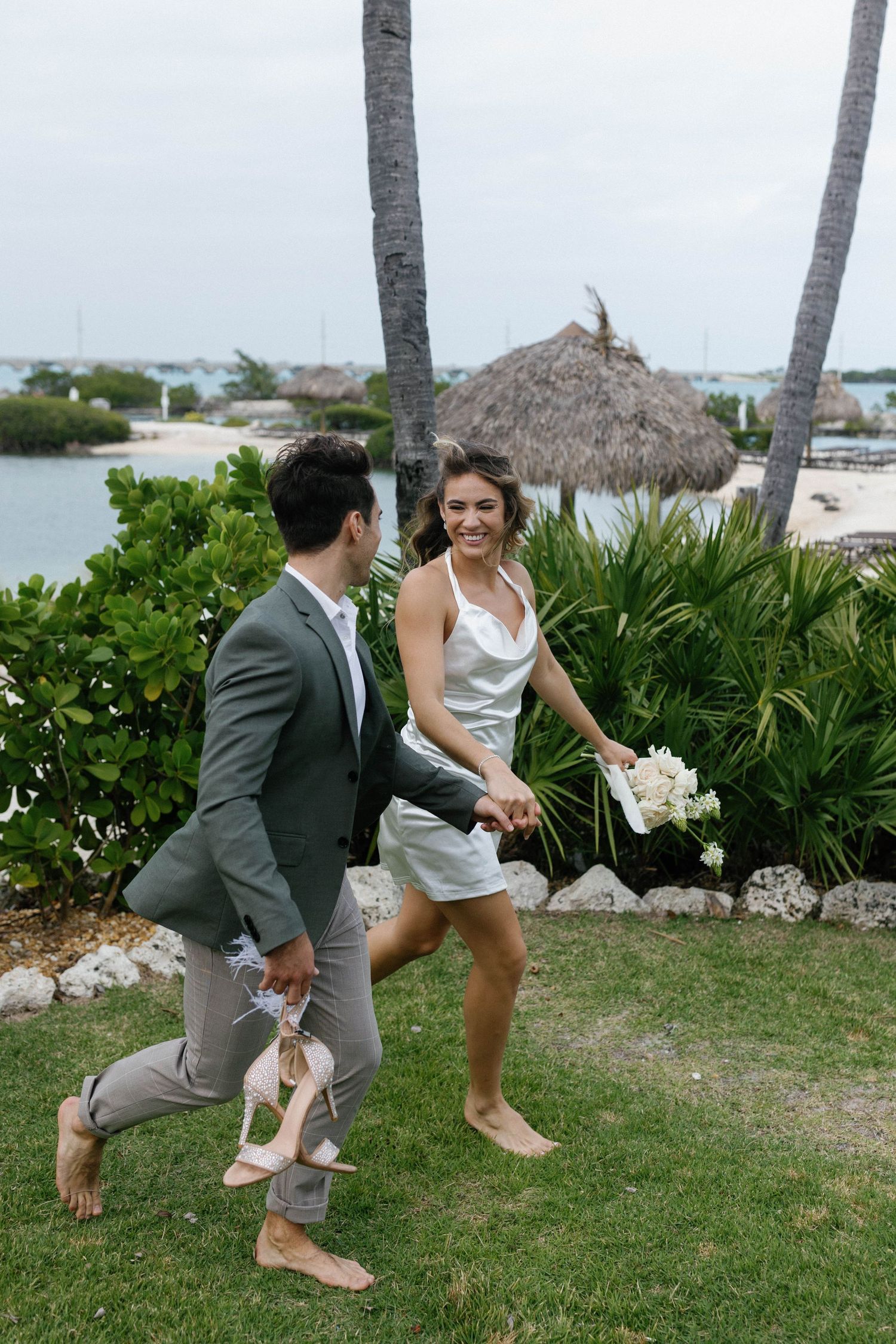 Playful couple running barefoot on grass near palm trees at tropical beach wedding.