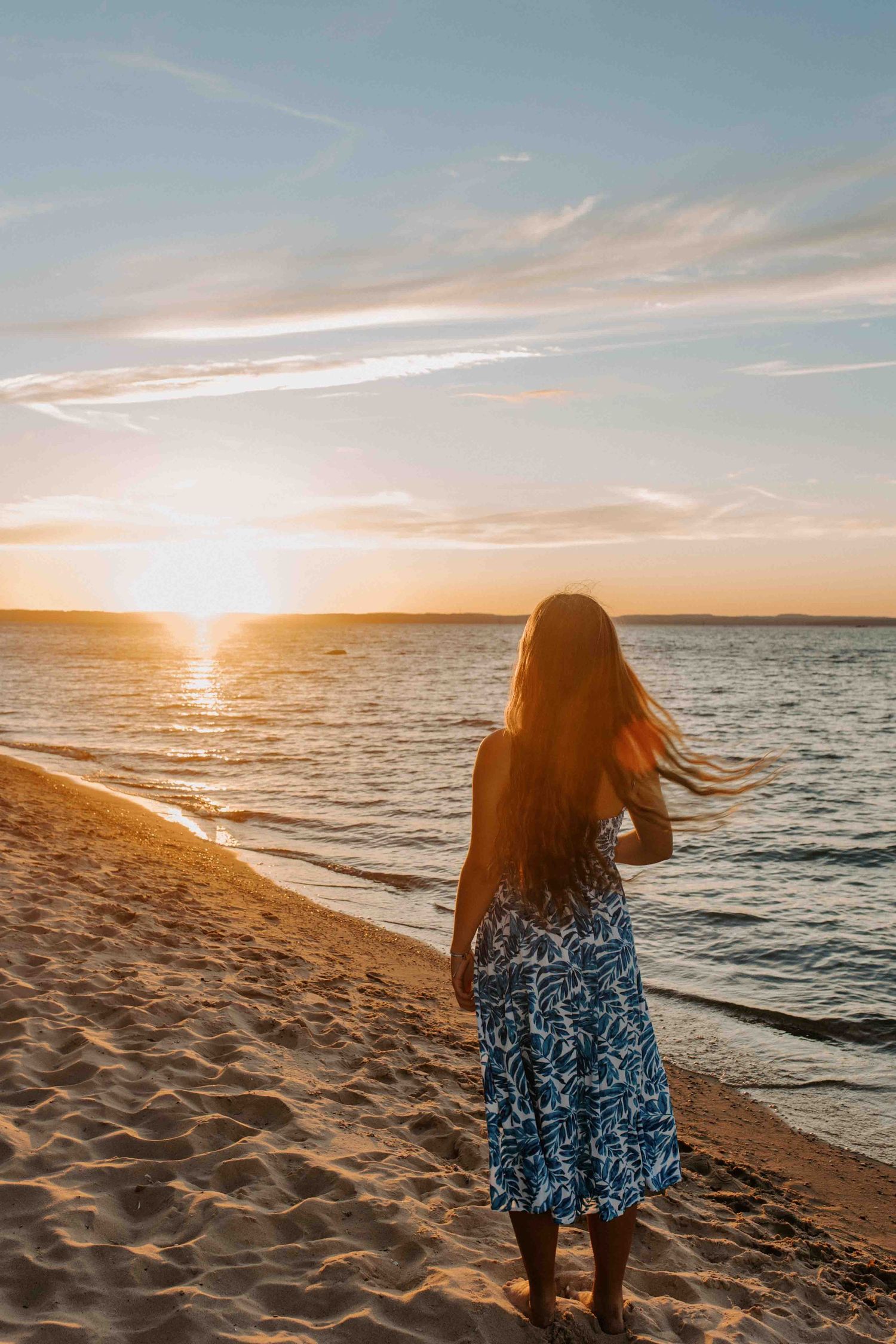 Person in a blue patterned dress walks along a sandy beach during sunset.