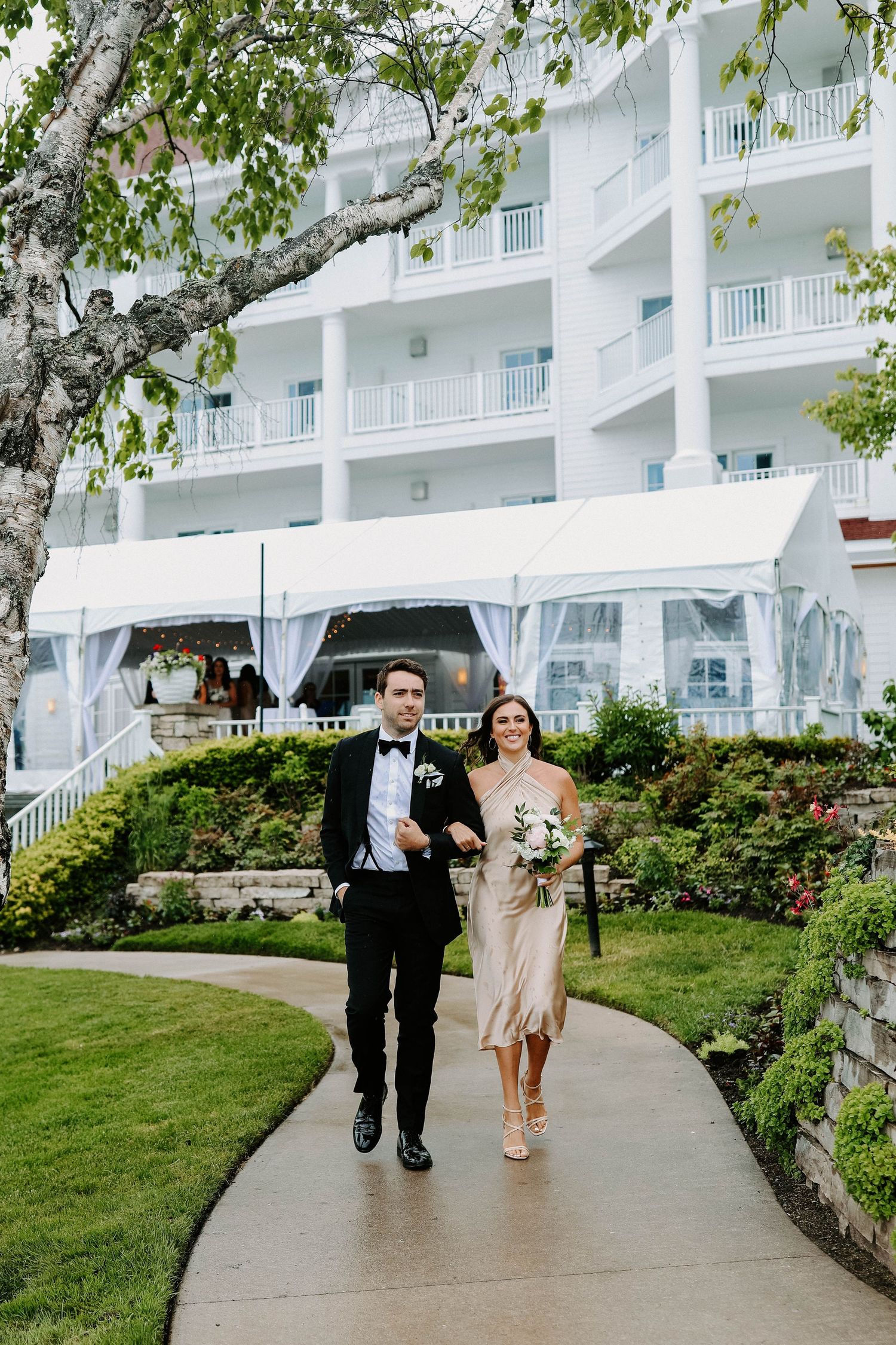 Wedding party members walk along a winding path with white resort building and green landscaping in background.