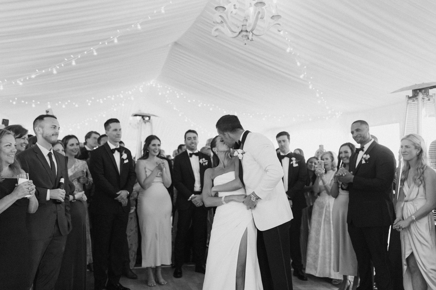 Wedding reception guests gathered on dance floor under white tent with twinkling lights.