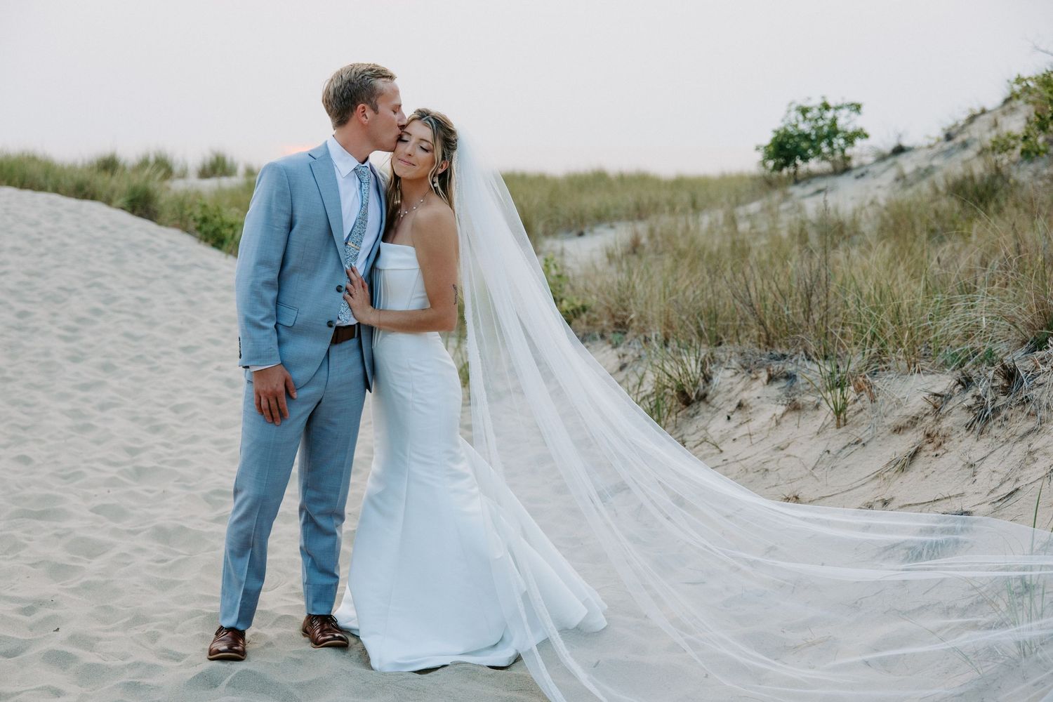 Couple in wedding attire shares tender moments on sandy beach with flowing veil in the breeze.