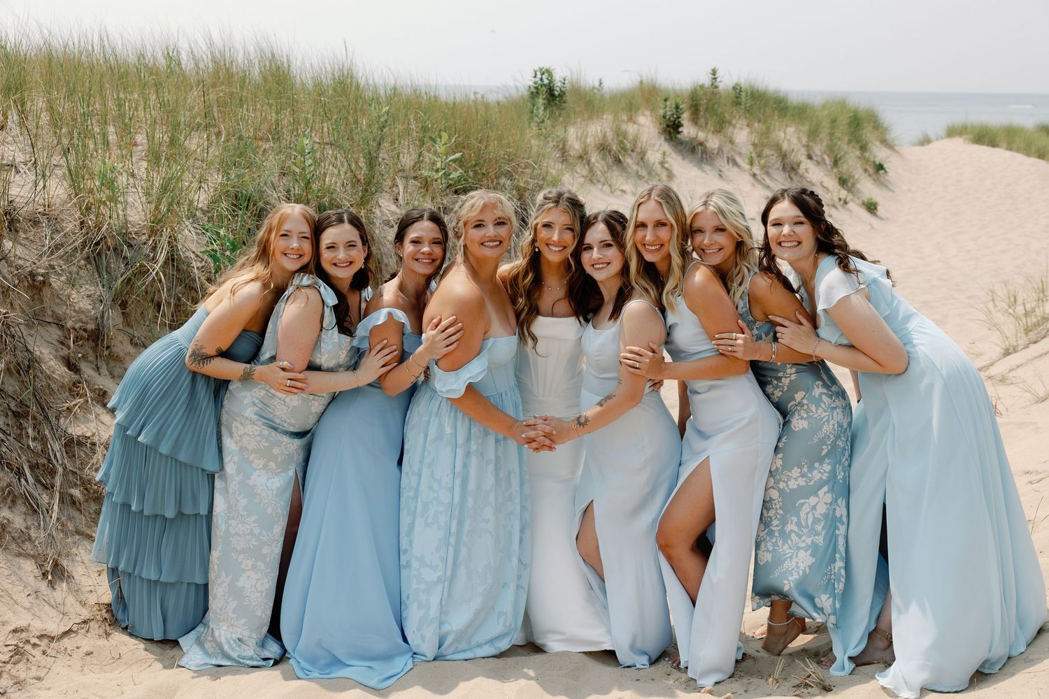 A wedding party in coordinated blue and white dresses poses together on a scenic beach.
