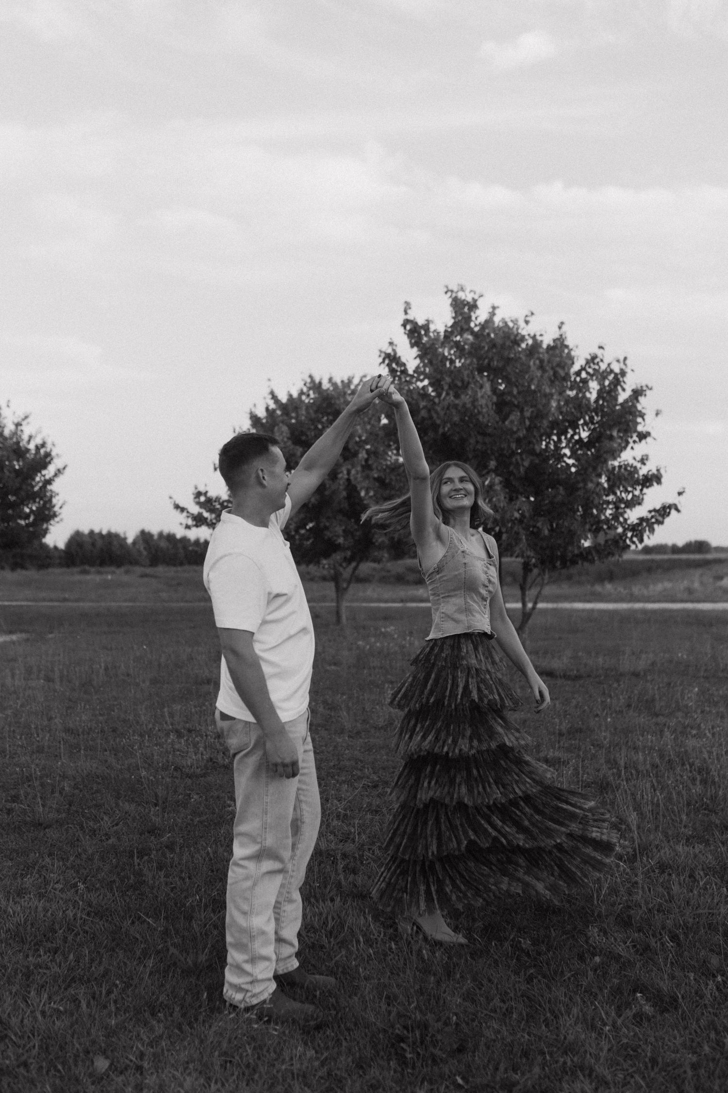 A black and white photo captures a dancing couple in an open field with trees.