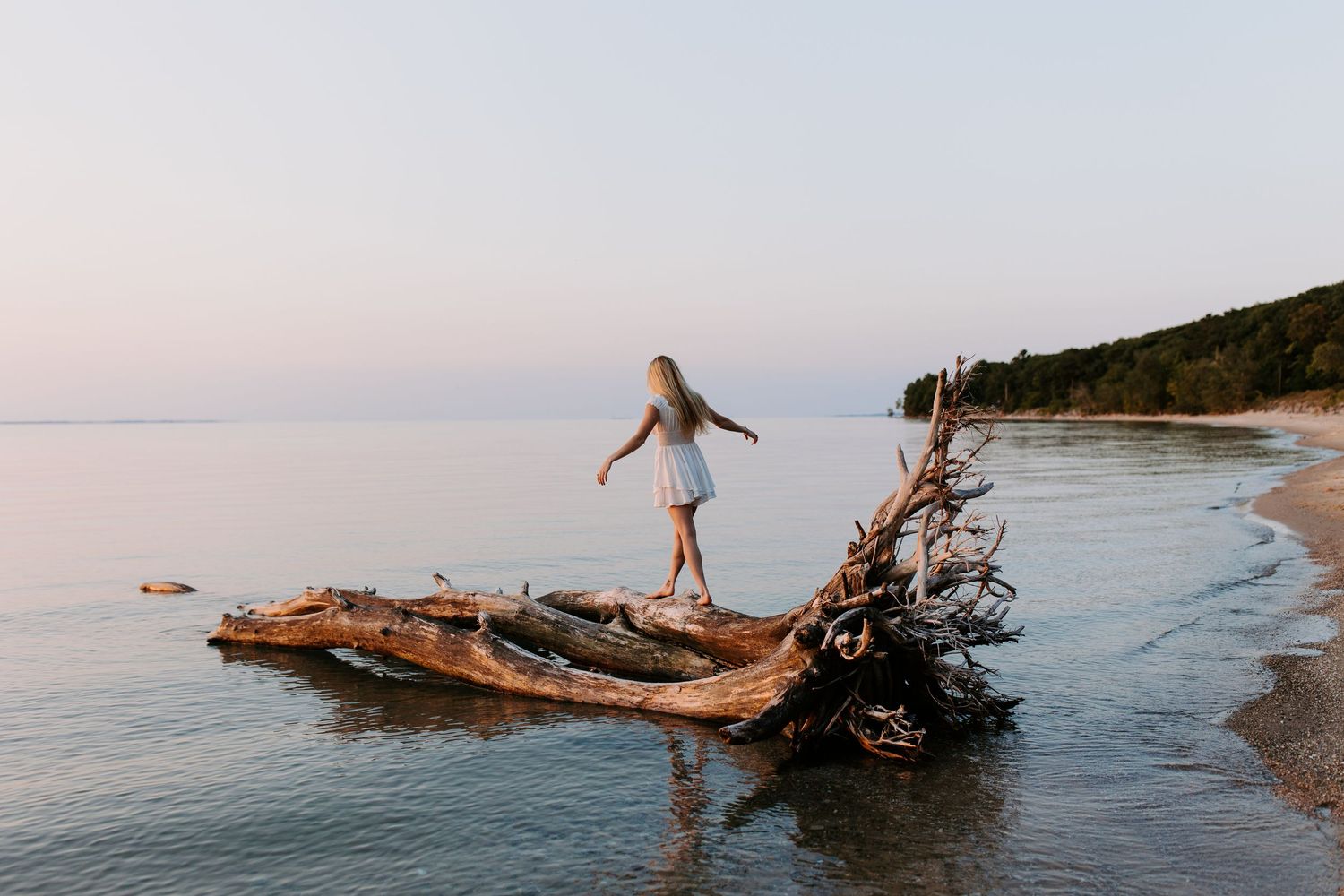 Person balancing on driftwood log at beach during sunset.