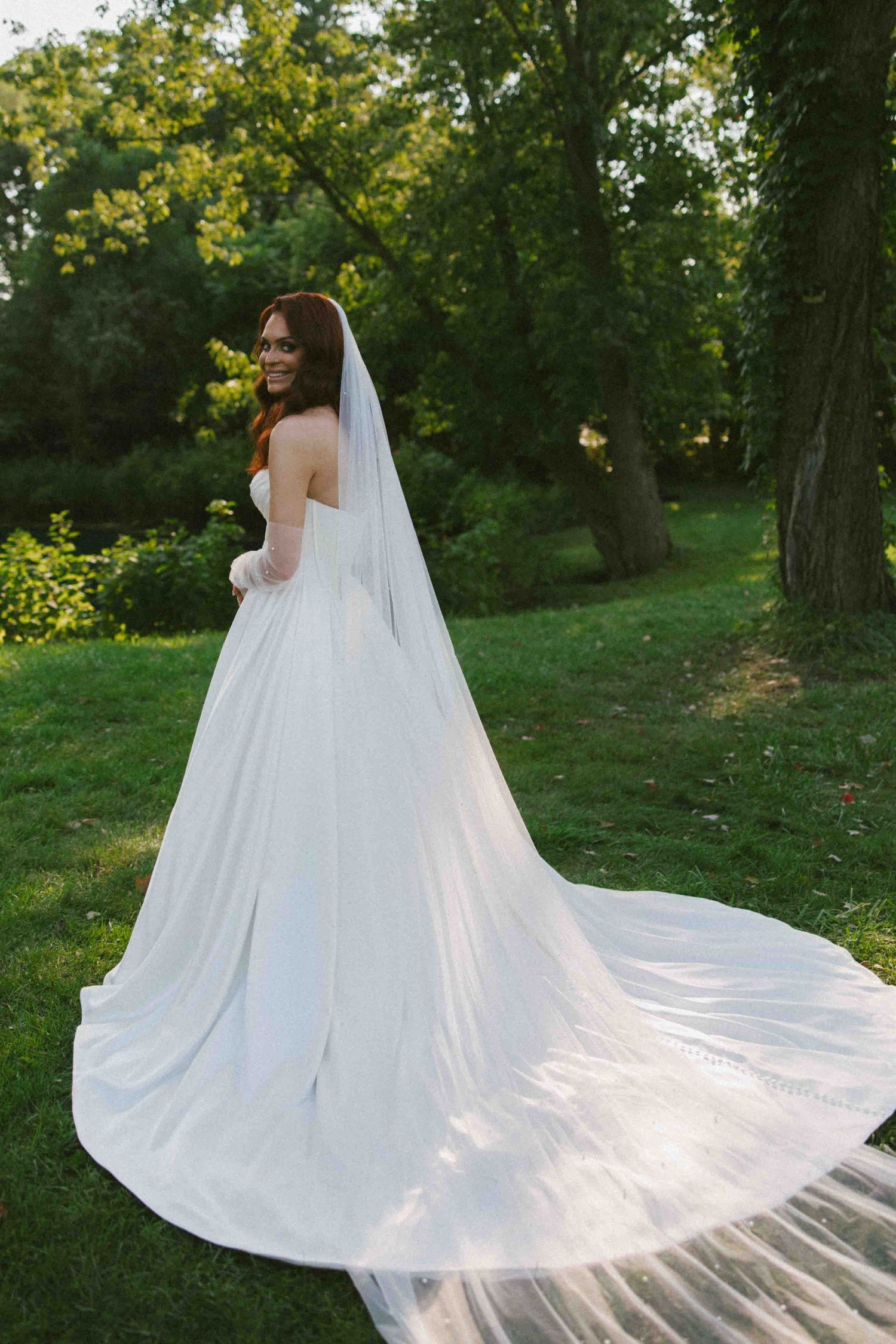 Flowing white wedding dress train spreads across green grass in outdoor setting.
