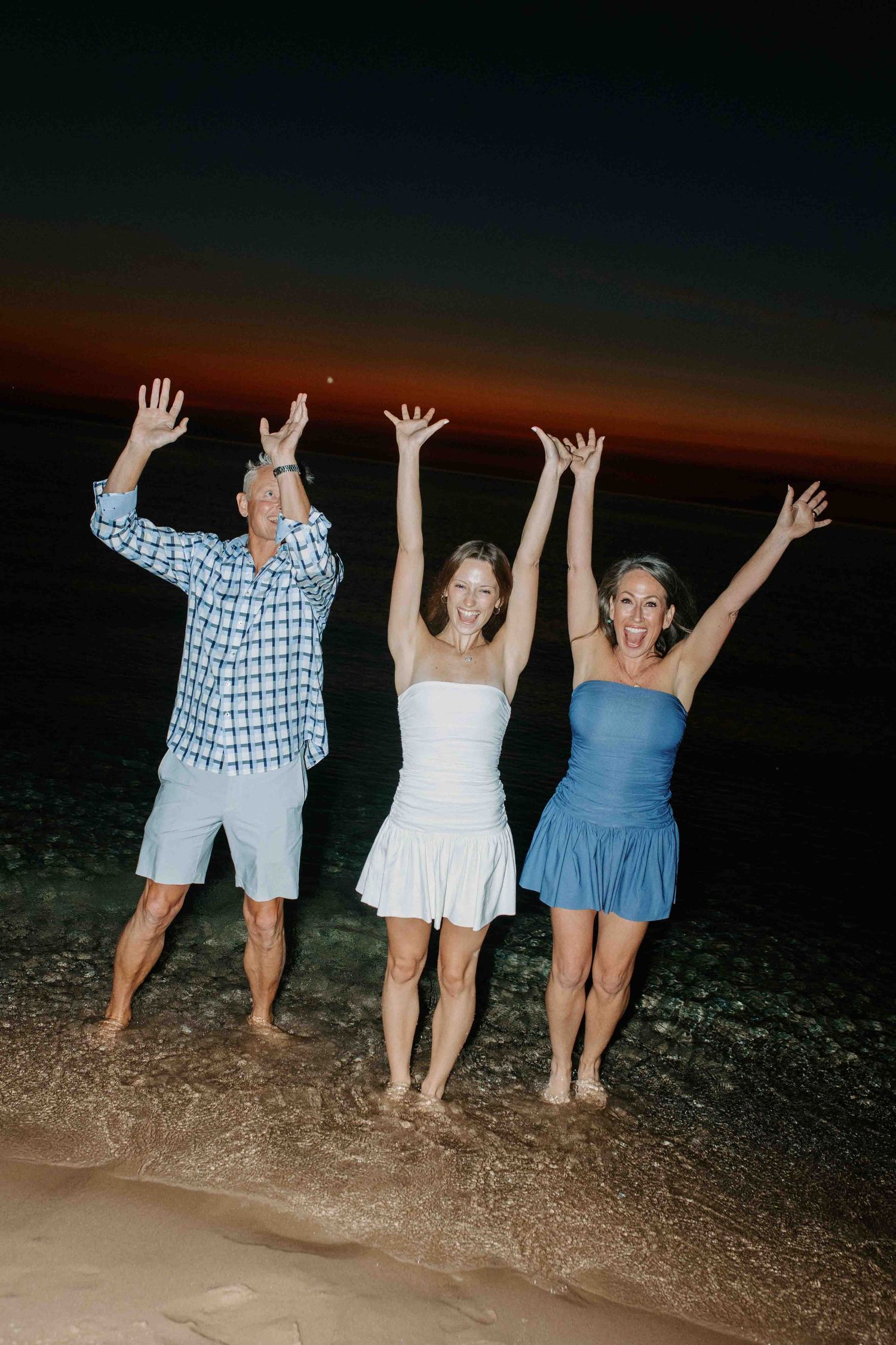 Four people raise their arms joyfully while standing in the ocean waves at sunset.