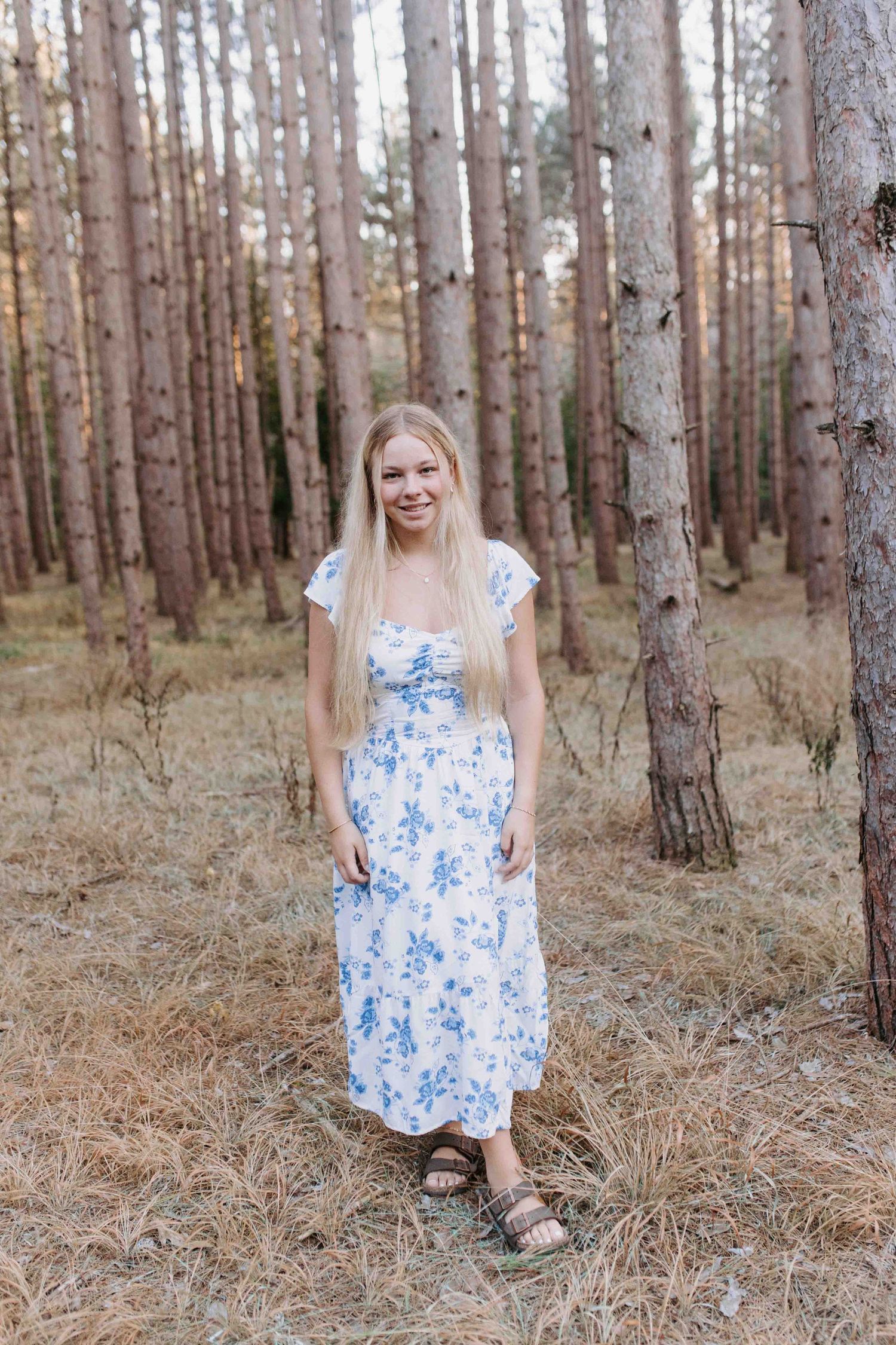 Standing among tall pine trees in a blue and white floral dress.