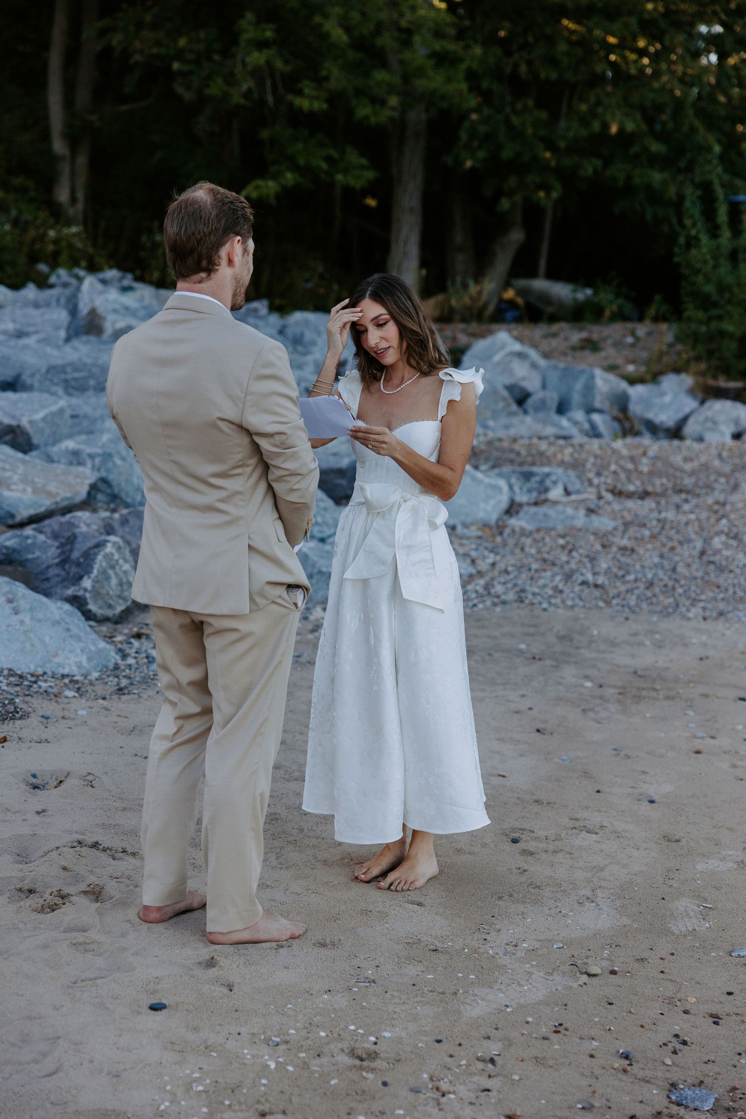 Couple in wedding attire walking barefoot on rocky beach during sunset photoshoot.