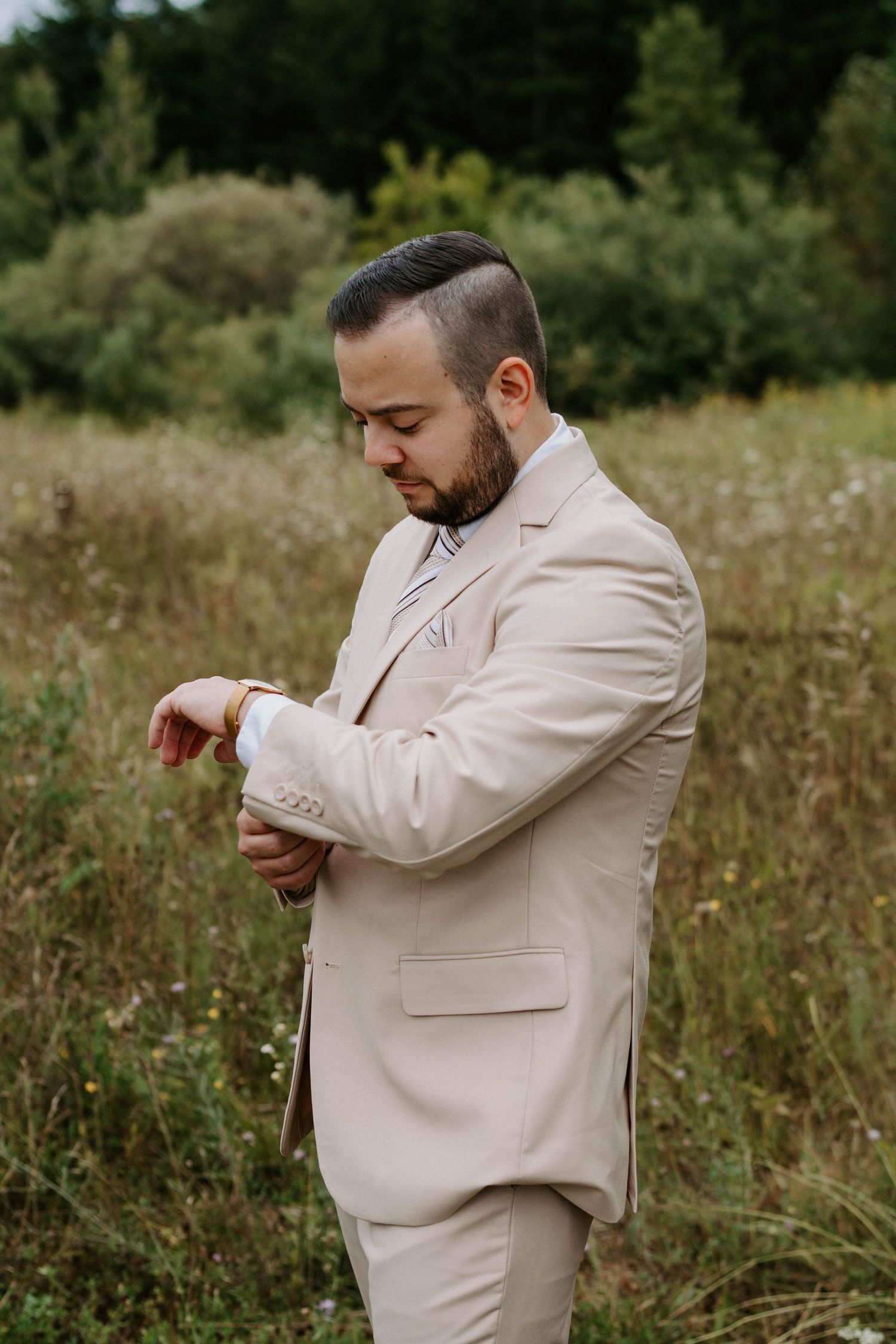 Groom in beige suit checks his watch while standing in a wildflower field.