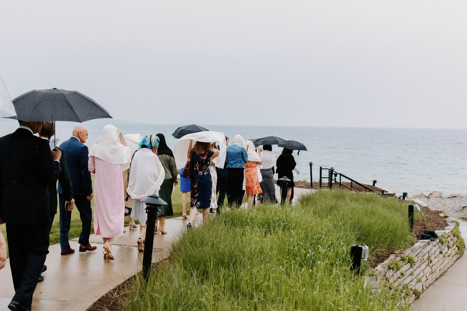 Wedding guests with umbrellas gathered on a cloudy beach for ceremony.