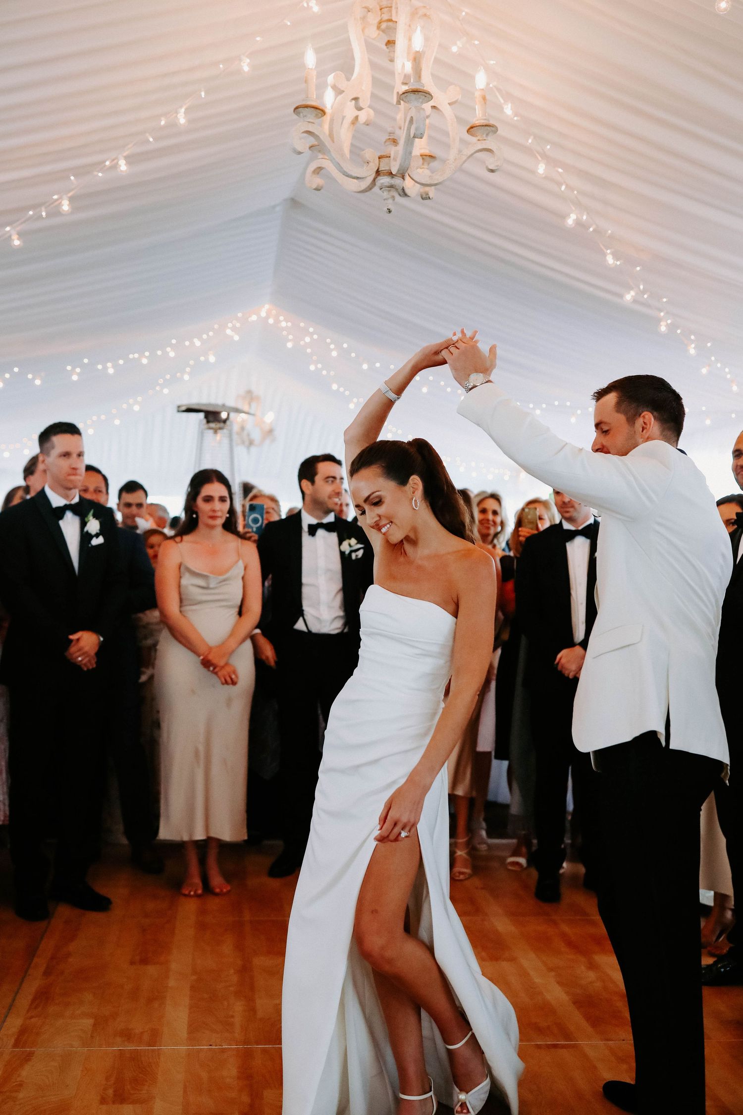 Couples dancing at an elegant wedding reception under white tent with twinkling lights and chandeliers.