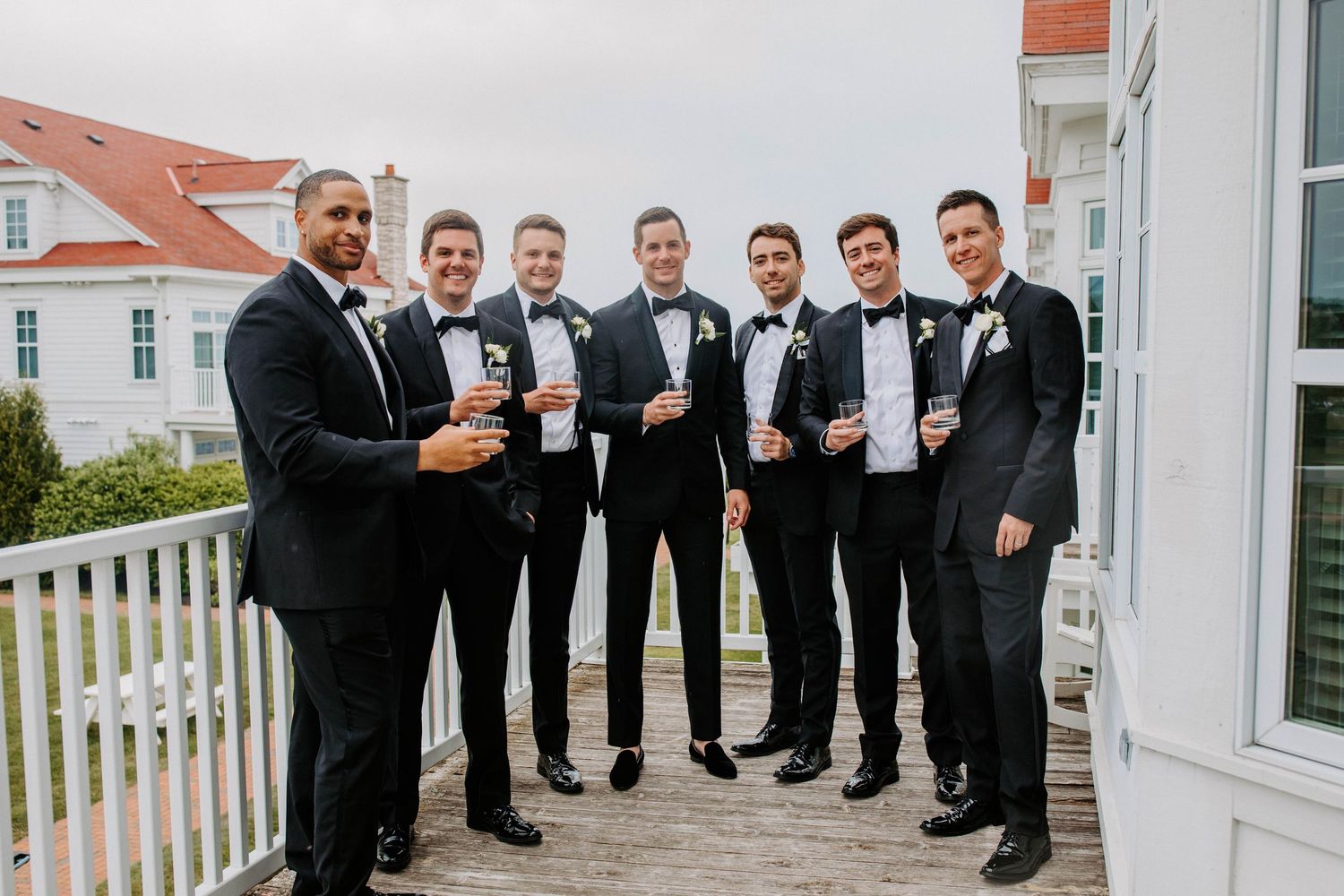Wedding party in formal black attire poses on a white porch with ocean views.