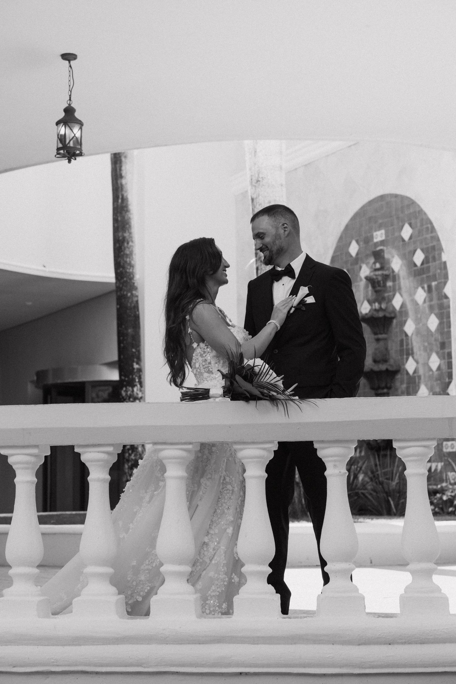 Black and white photo of couple sharing intimate moment on balcony with ornate railings.