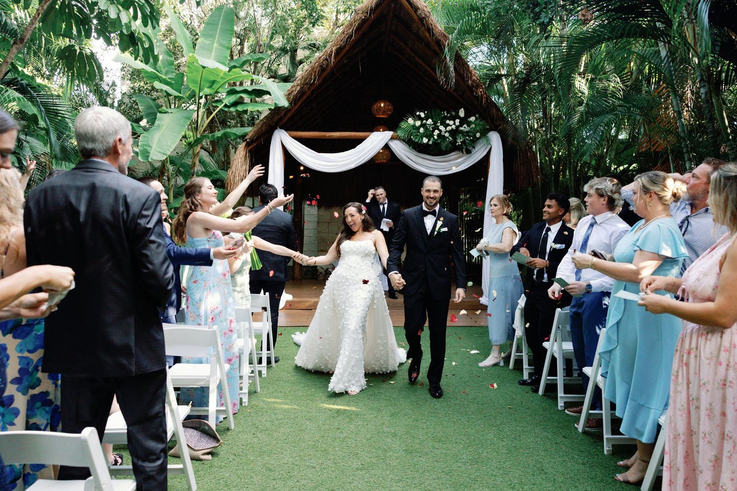 Wedding guests seated in white chairs at an outdoor garden ceremony with decorative white fabric draping overhead.