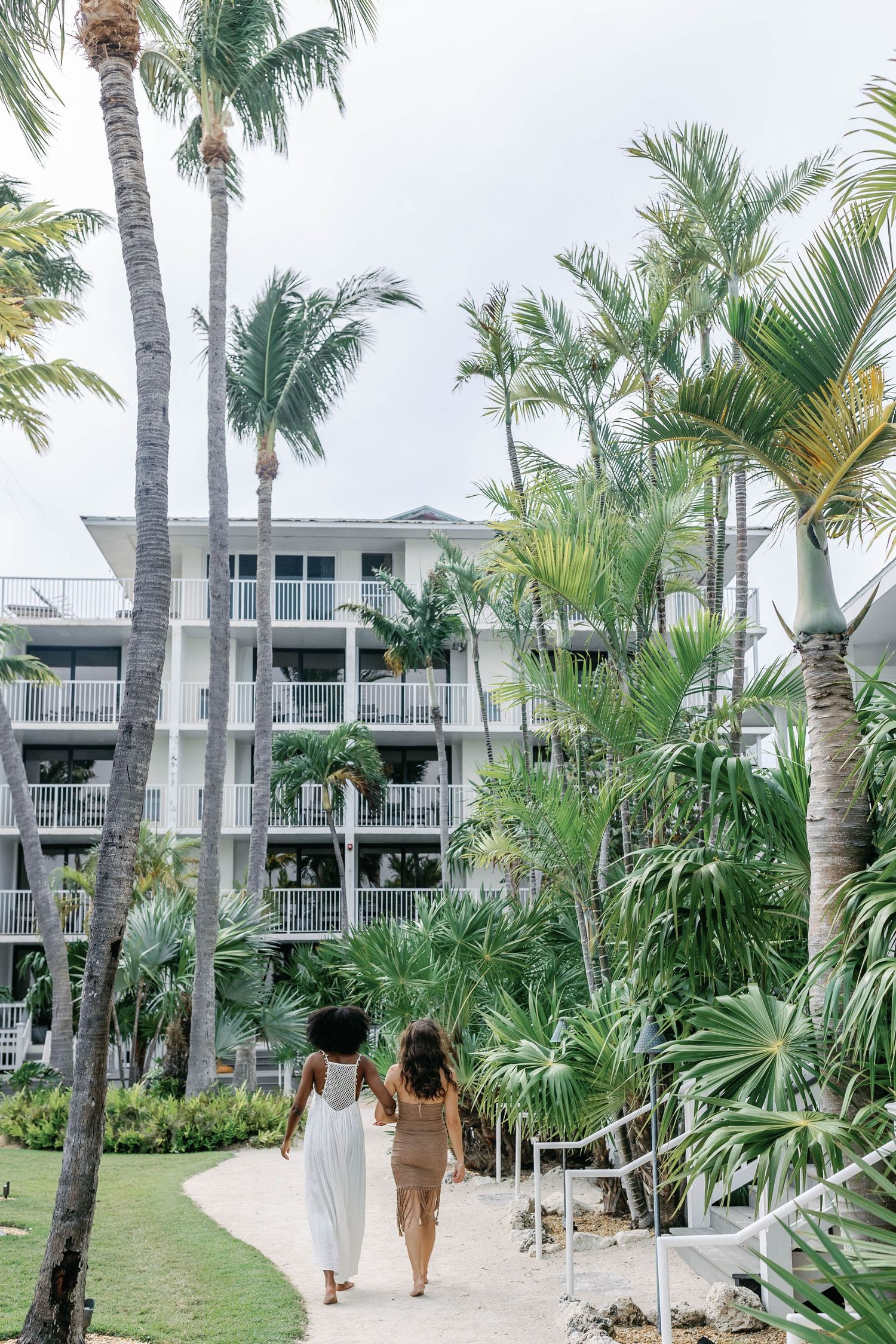 Palm trees line a walkway leading to modern white beachfront condominiums with tropical landscaping and balconies.