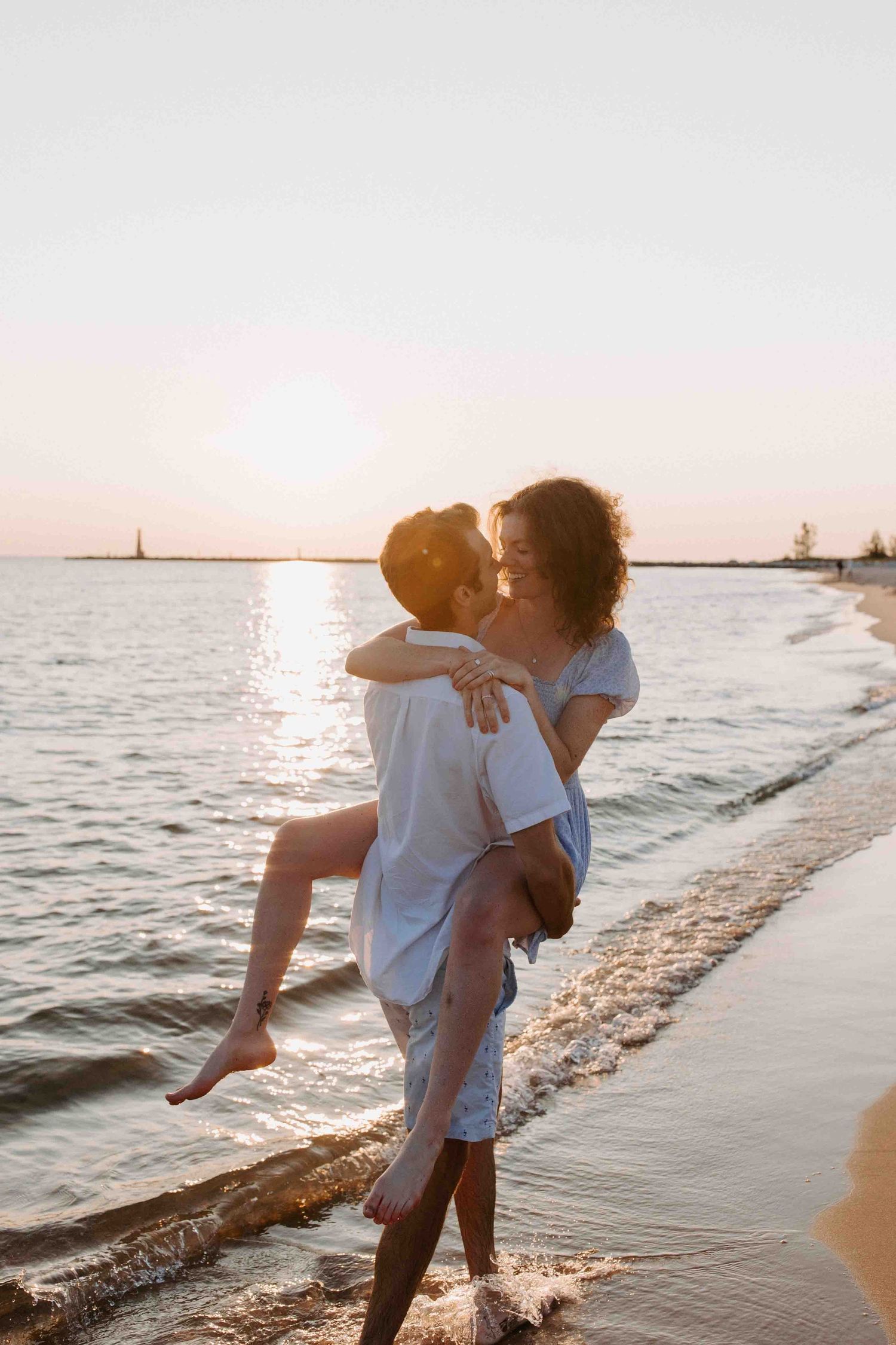 A romantic couple embraces on a sandy beach at sunset with gentle waves in the background.