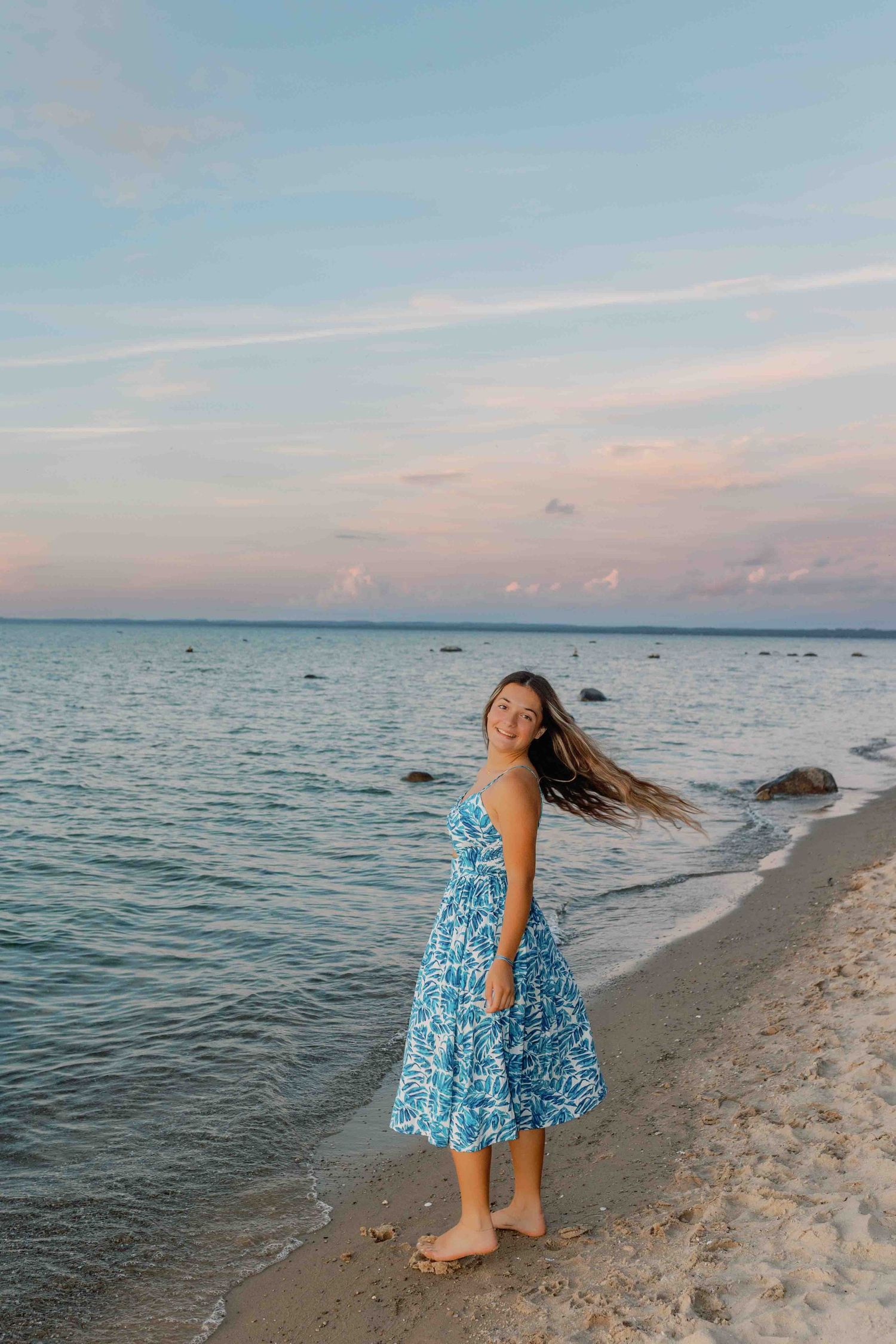 Someone in a flowing blue dress dances playfully on a rocky beach shoreline during golden hour.