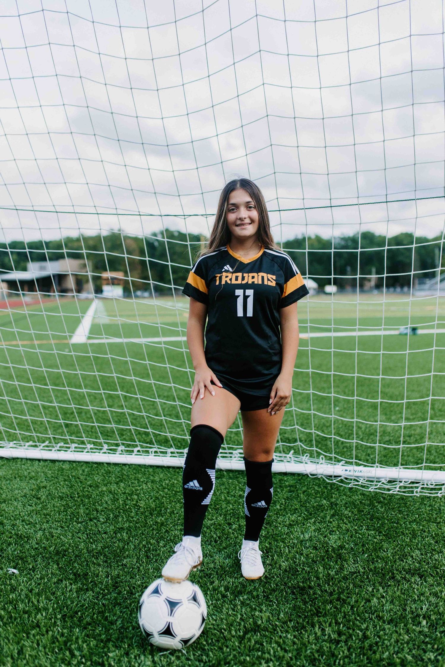Soccer player in black Trojans uniform poses with ball on field by goal net.