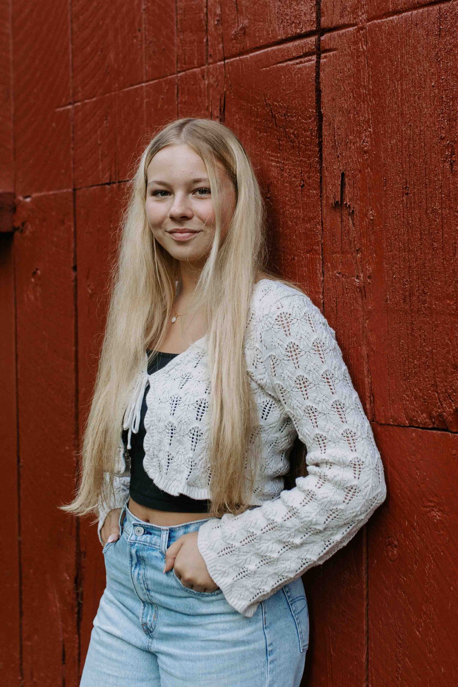 Fashion poses against a rustic red barn wall in a white knit sweater and light denim jeans.