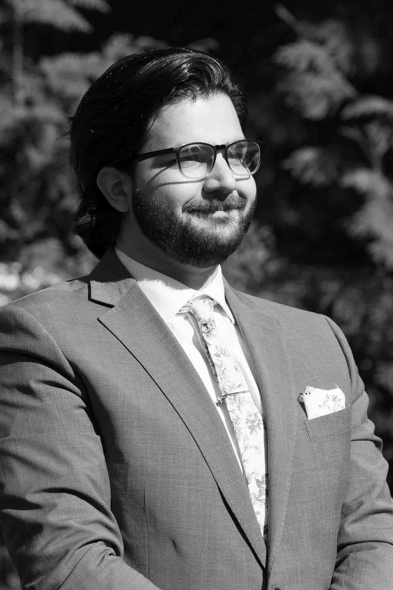 Groom in formal grey suit and patterned tie smiling during outdoor ceremony.