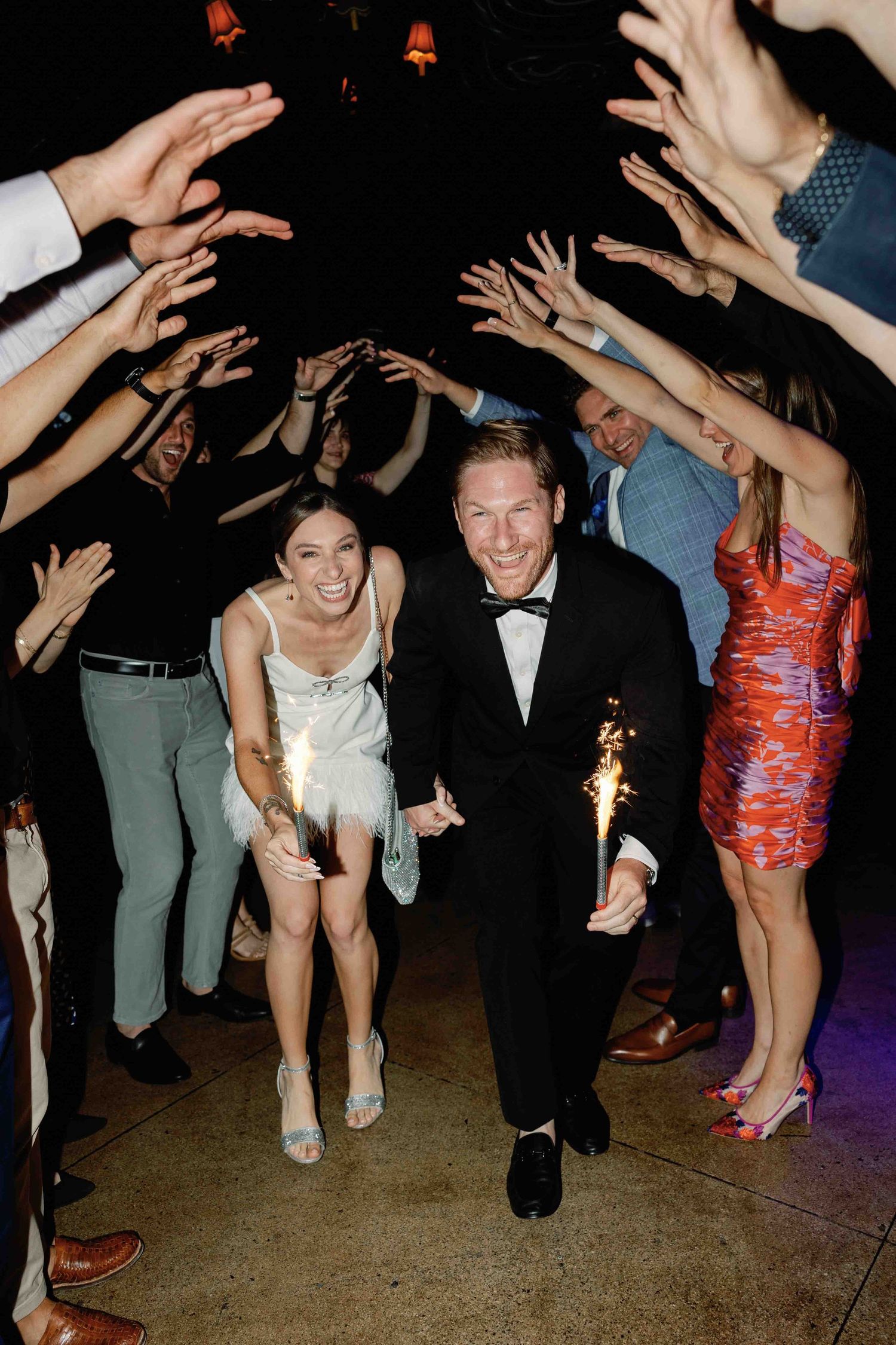 Wedding guests dance and celebrate on a dark dance floor with hands raised in the air.