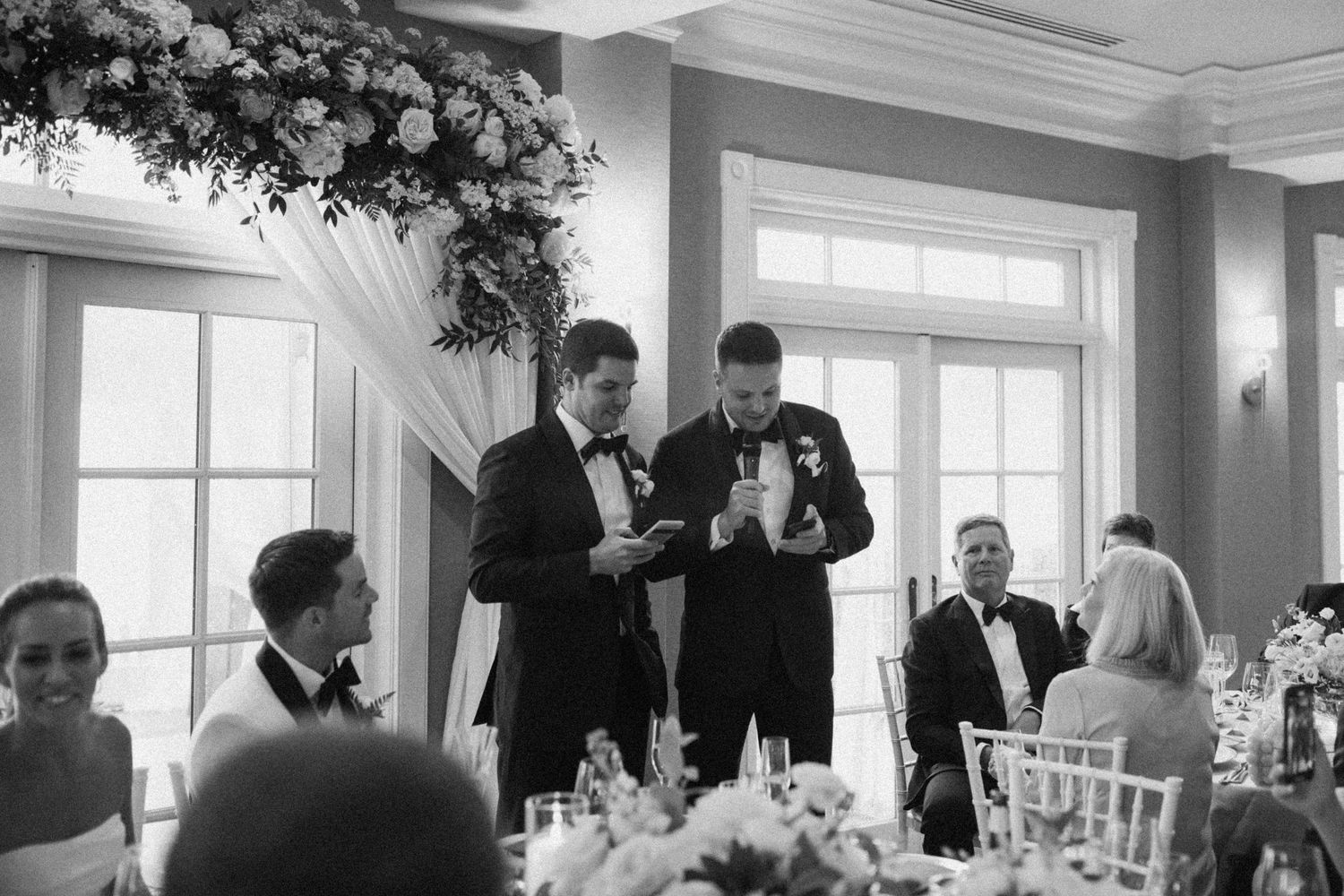 Black and white panoramic view of a wedding reception hall with guests dining at decorated tables.
