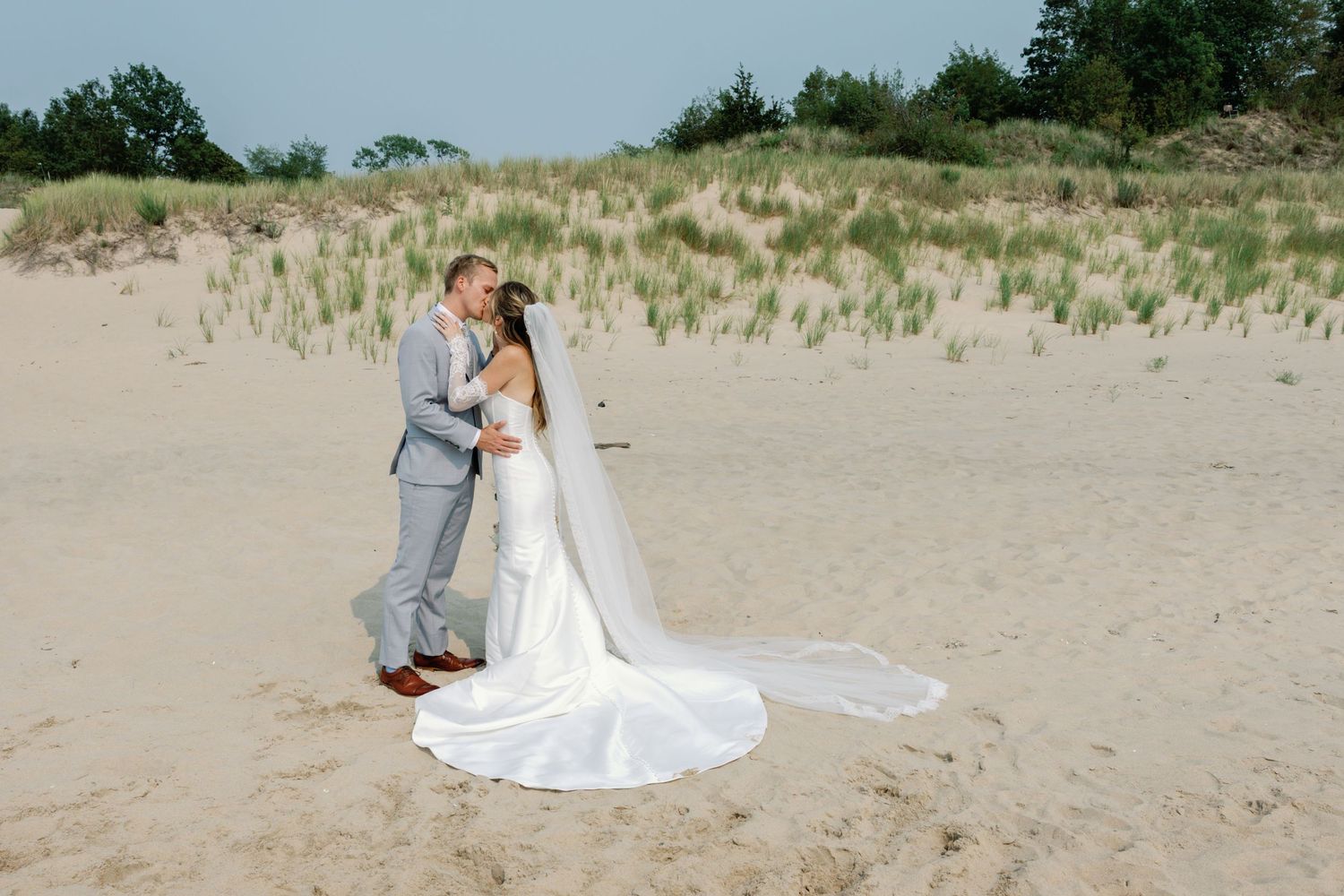 A couple in wedding attire poses together on a sandy beach with dunes and grass in the background.