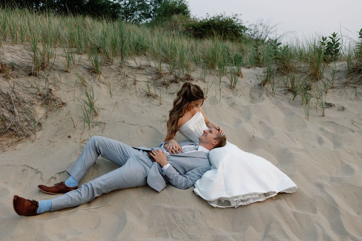 Playful wedding photos of couple lying in beach sand wearing formal attire.