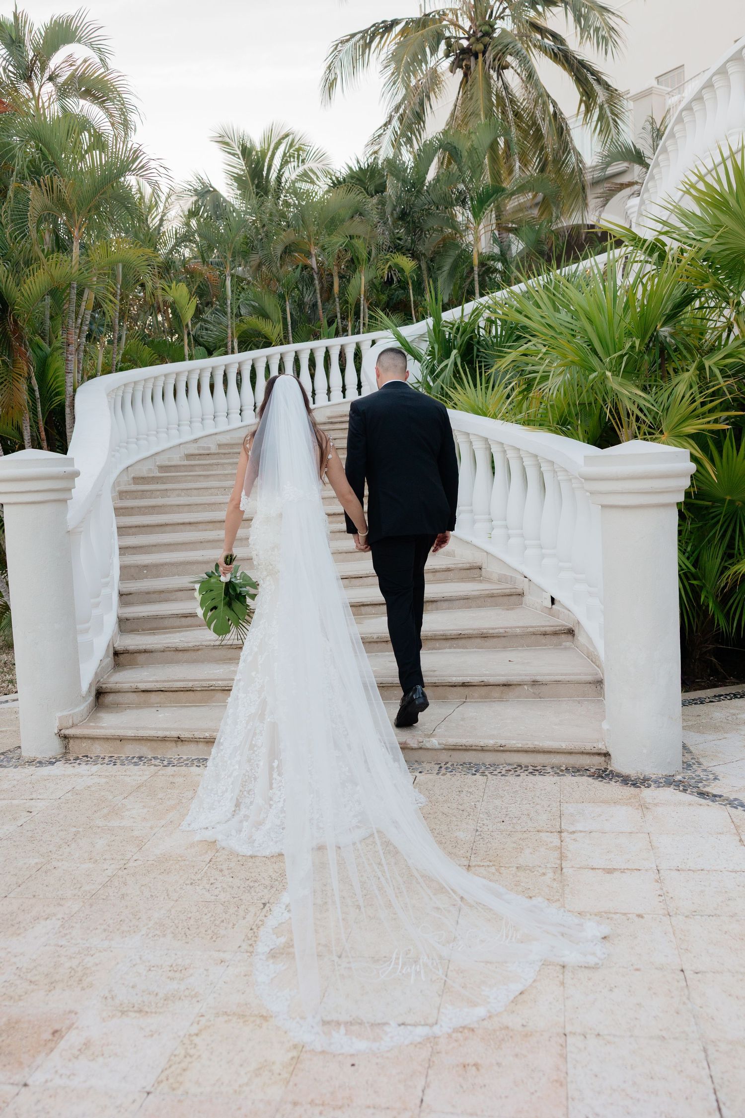A bride and groom walk up elegant white stairs surrounded by tropical palm trees at Dreams Tulum Resort and Spa